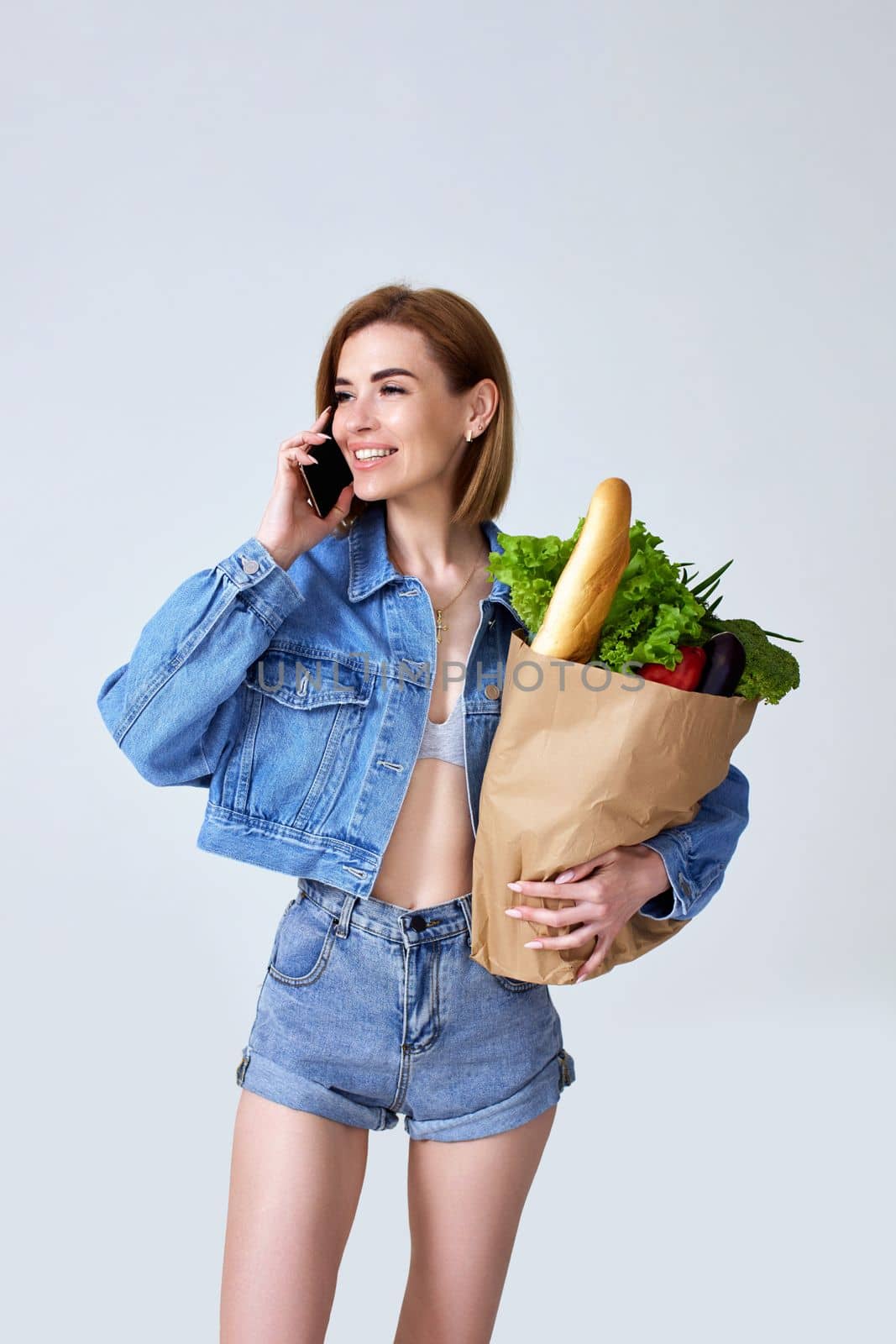 vegetarian woman holding shopping eco-friendly paper bag full of groceries and using smartphone on studio gray background.