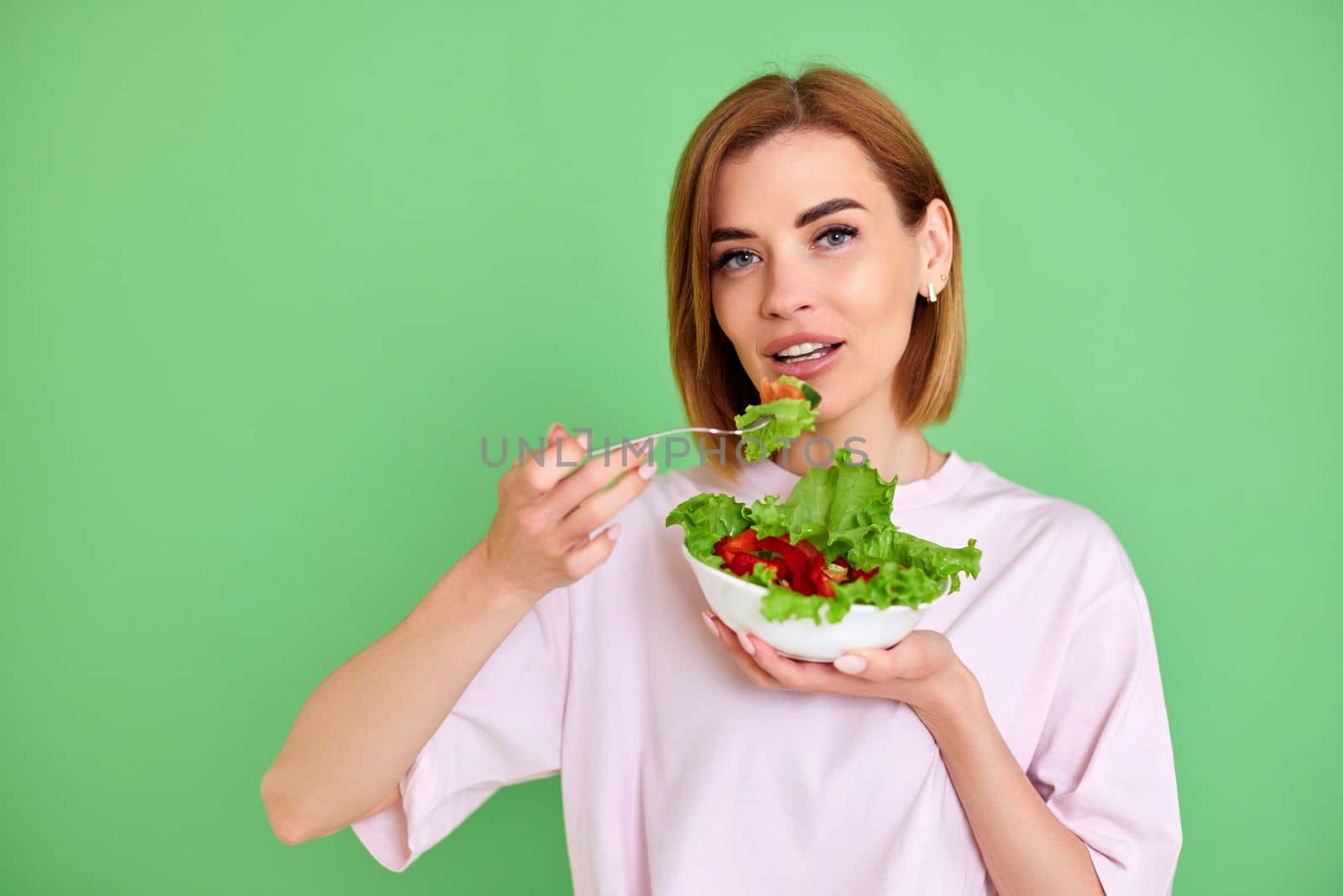 Cheerful blonde woman eating fresh vegetable salad on green background