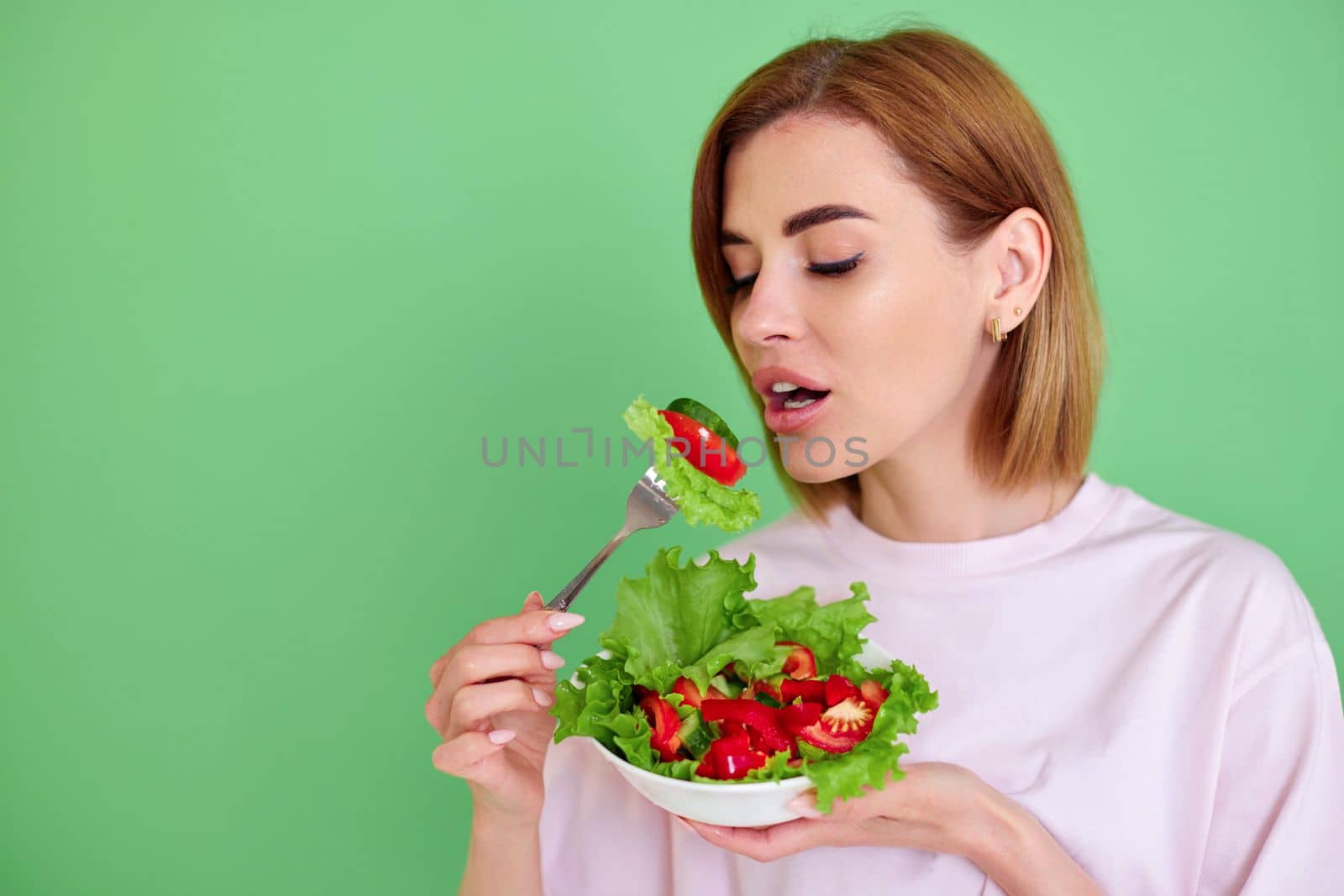 Cheerful healthy woman eating fresh vegetable salad on green background