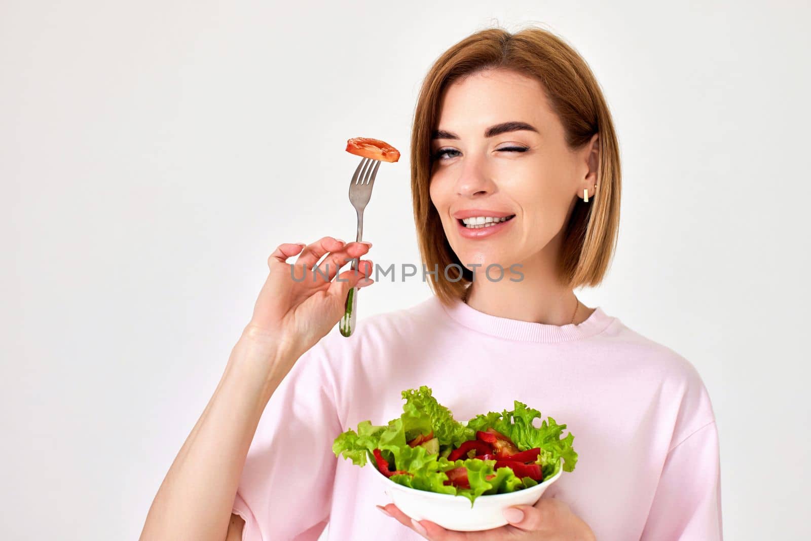 beautiful smiling blonde girl eating fresh vegetable salad and winking on white background