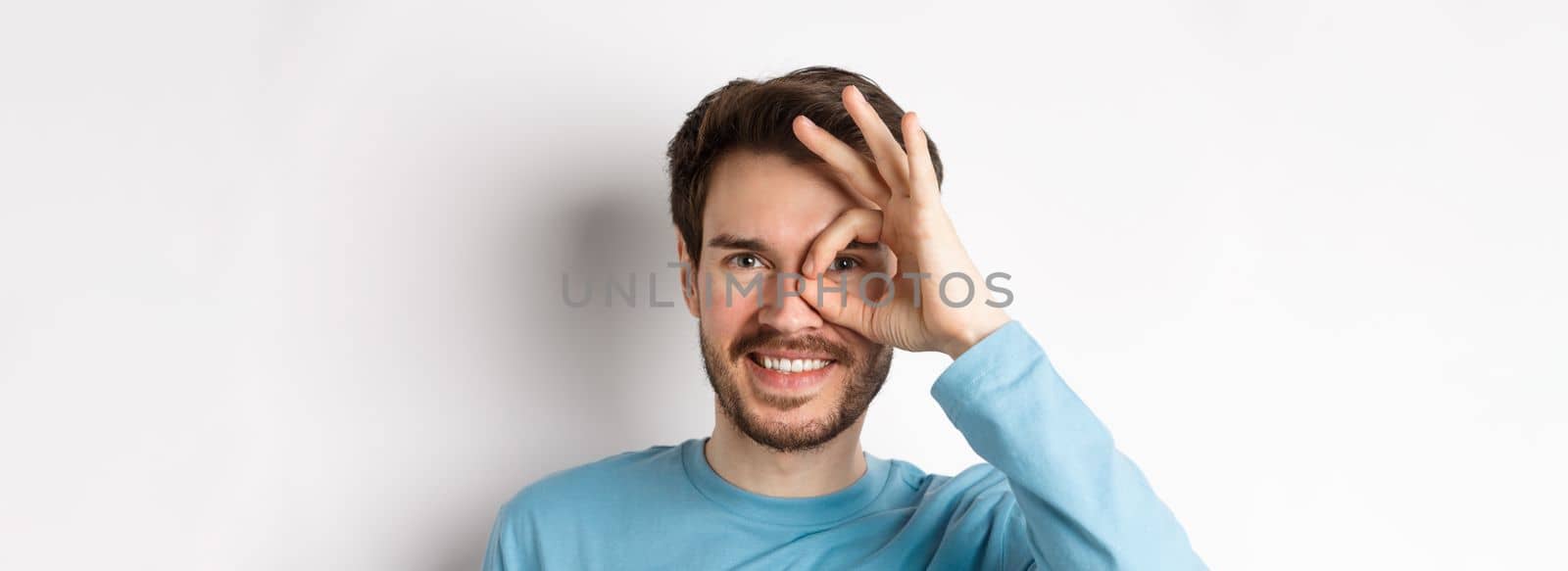 Close up of happy man looking through okay sign and smiling, approve and like something good, standing over white background.