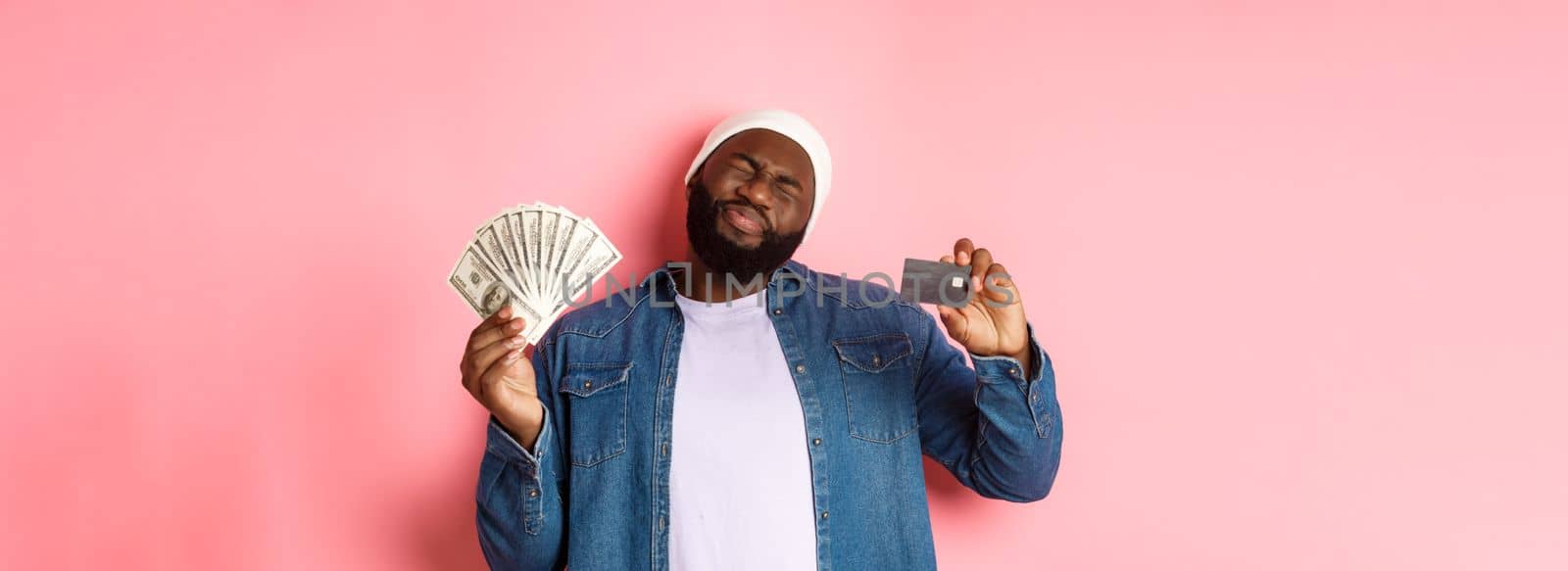 Shopping concept. Sad and whining Black guy showing credit card and dollars, grimacing reluctant, standing over pink background.