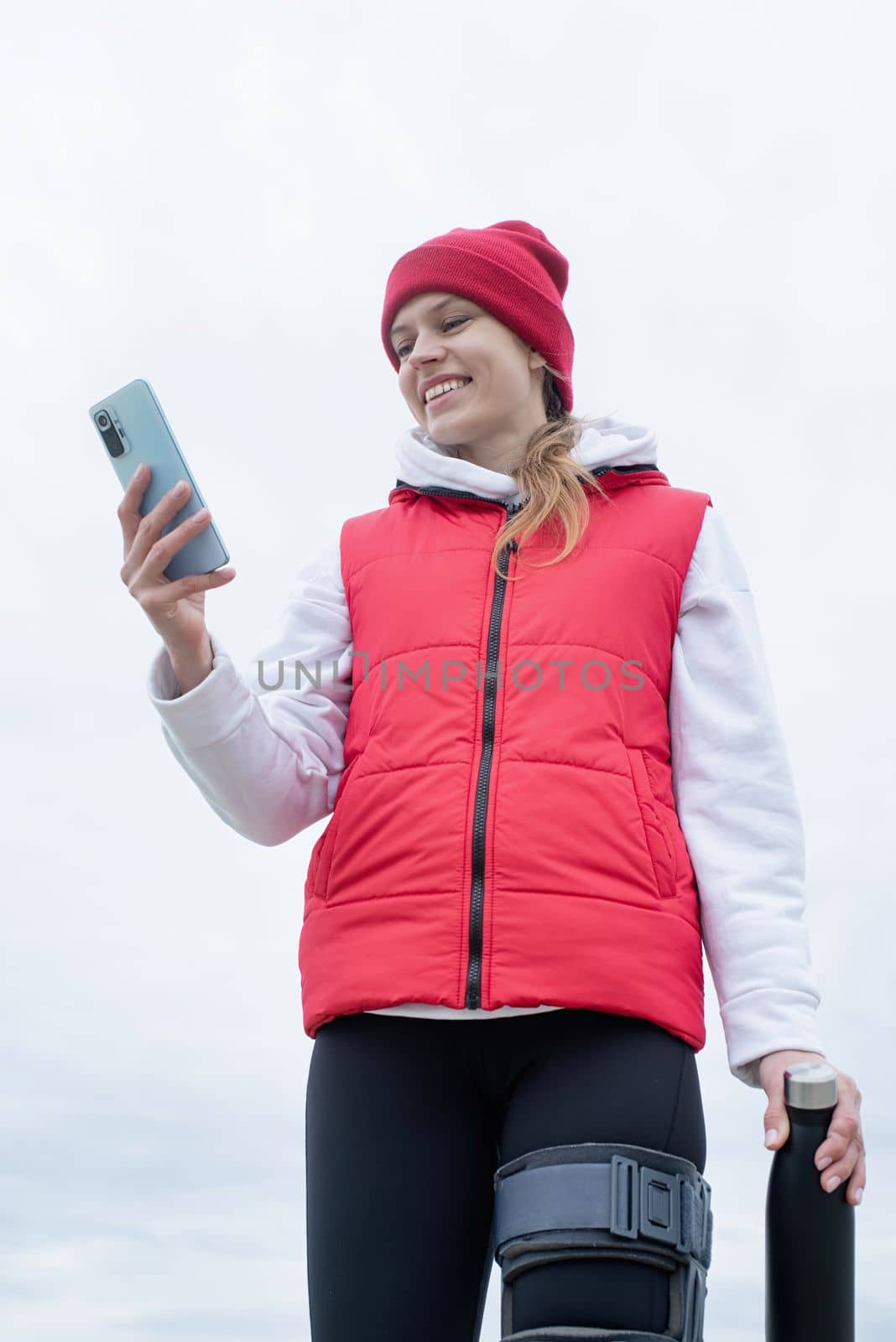 Woman wearing sport clothes and knee brace or orthosis after leg surgery walking in the park using smartphone. Medical and healthcare concept.