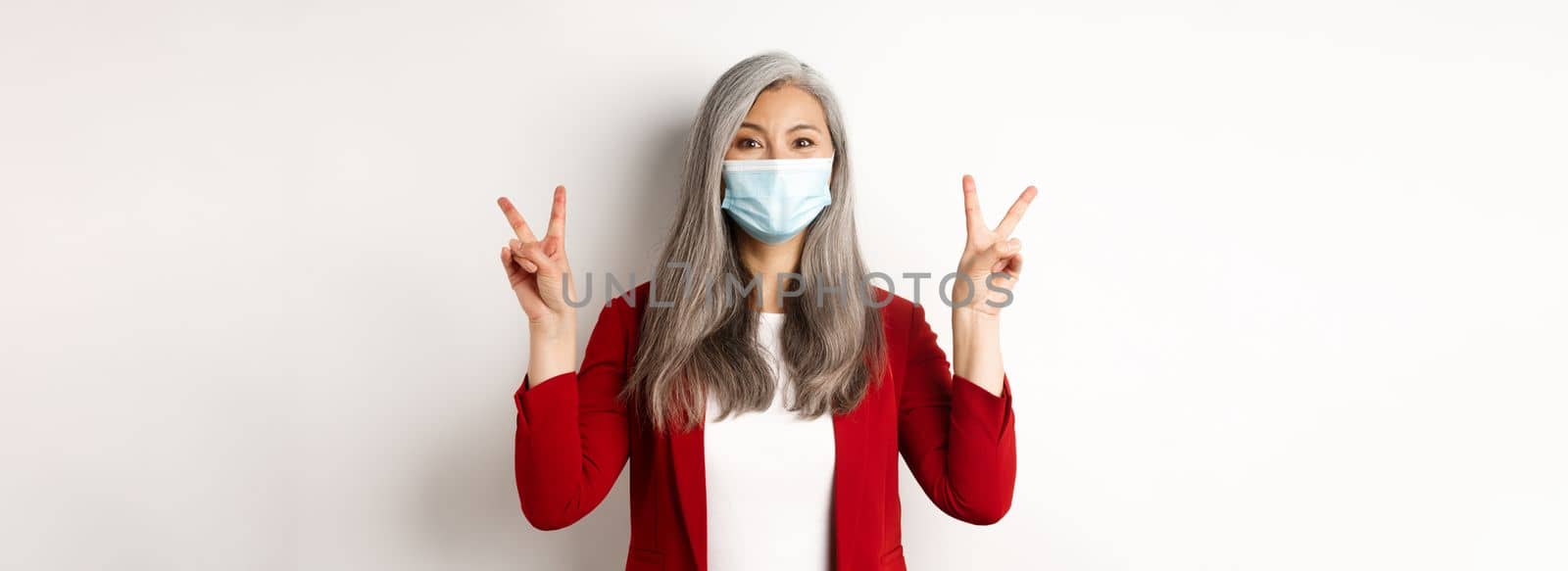 Coronavirus and business concept. Cheerful senior woman in face mask, showing victory or peace sign and smiling with eyes, white background.