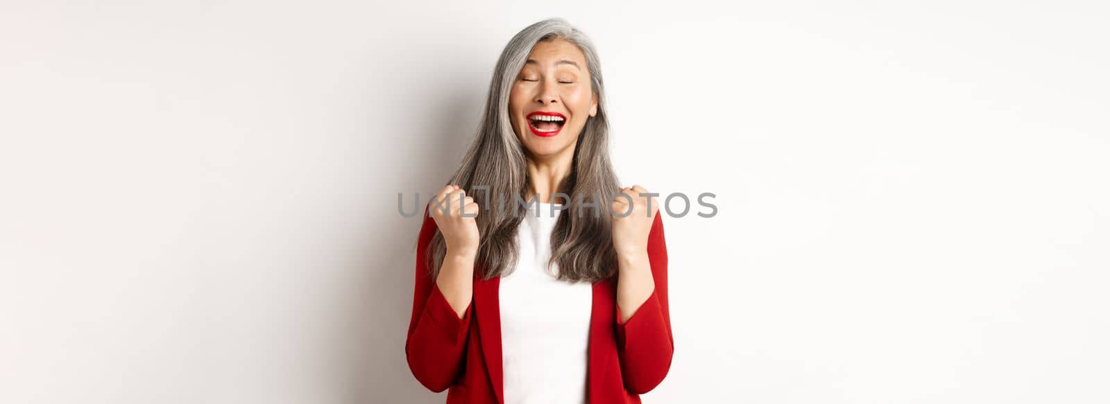 Relieved asian senior businesswoman making fist pump, saying yes and smiling satisfied, triumphing and winning, standing over white background.