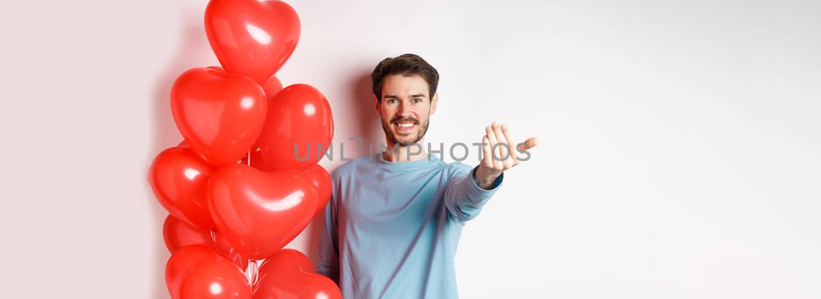 Smiling man beckon you to come closer, follow me gesture, taunting his lover move forward, have romantic surprise, standing near red balloon on valentines day, white background by Benzoix