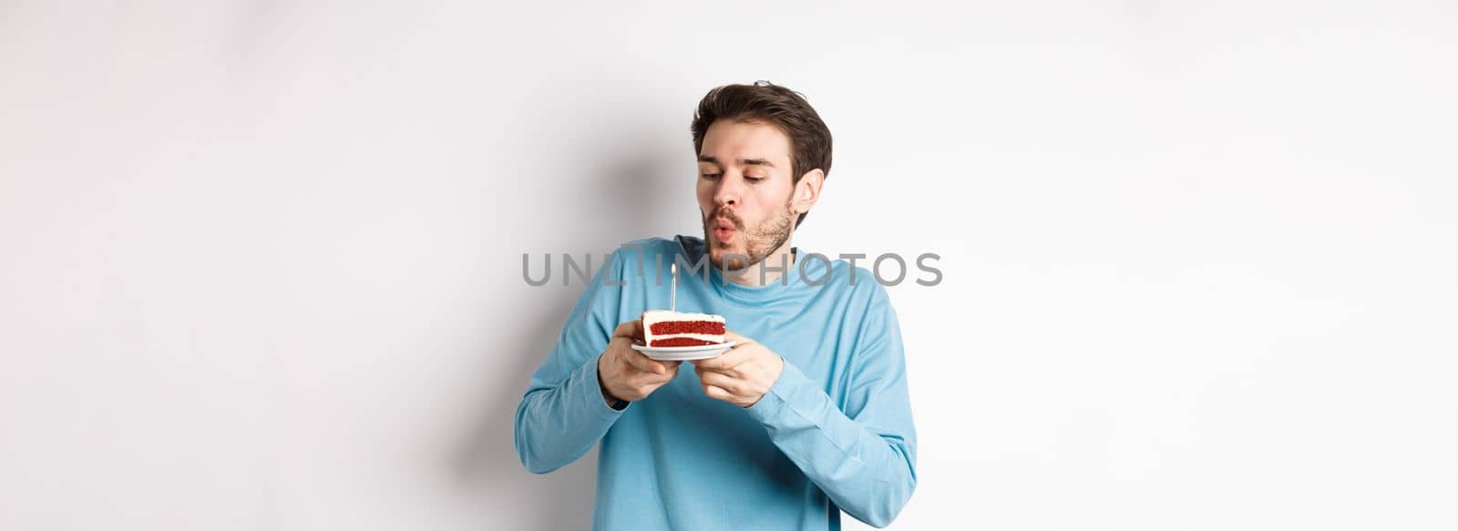 Celebration and holidays concept. Happy young man blowing candle on birthday cake, making wish on bday, standing over white background.