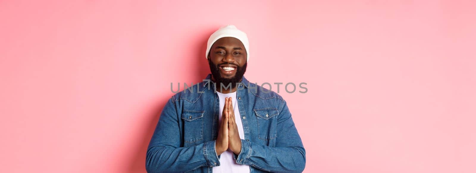 Happy smiling Black man saying thank you, holding hands in pray or namaste gesture, standing grateful against pink background.