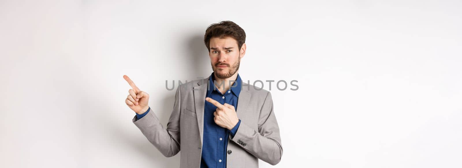 Showing bad example. Skeptical businessman look displeased and frowning while pointing at upper right corner banner, standing against white background.