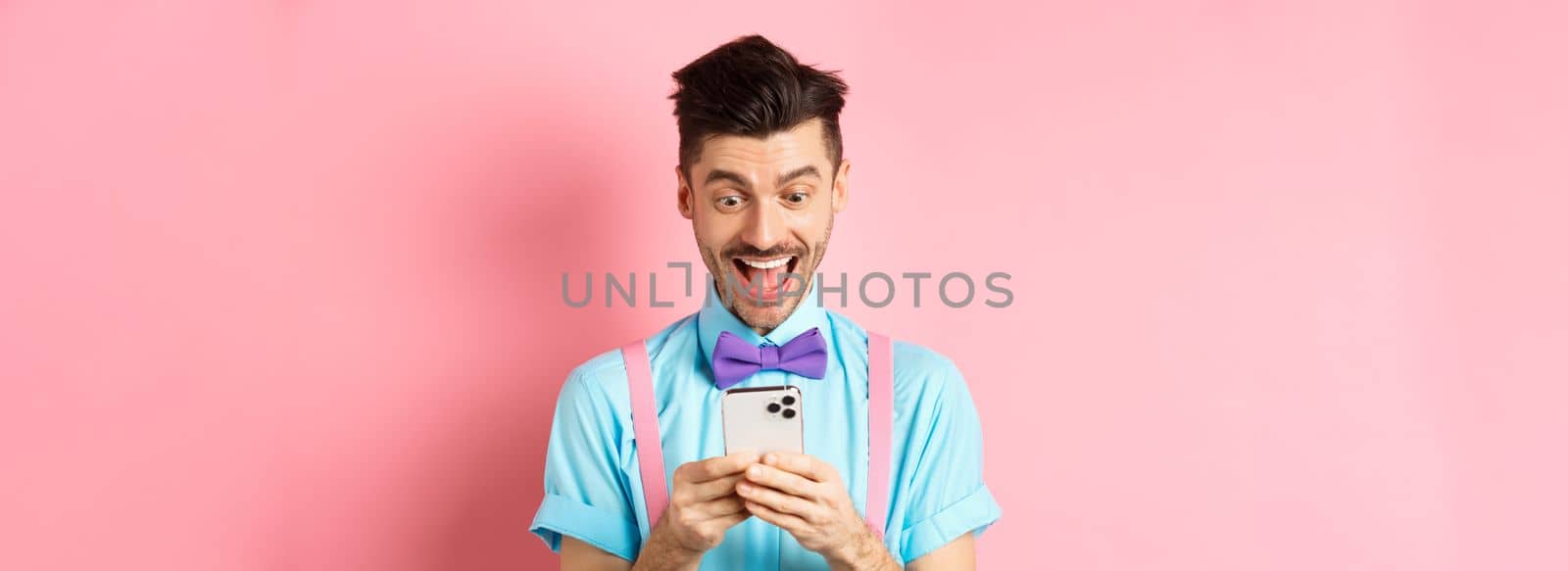 Technology concept. Cheerful young guy winning online prize, staring happy at smartphone screen, reading message amazed, standing on pink background.