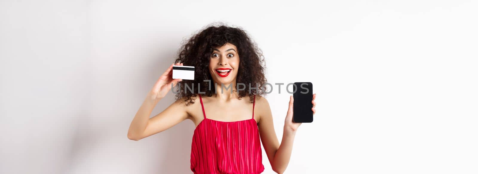 Online shopping concept. Excited curly-haired woman in red dress showing empty smartphone screen and plastic credit card, smiling happy at camera, standing on white background.