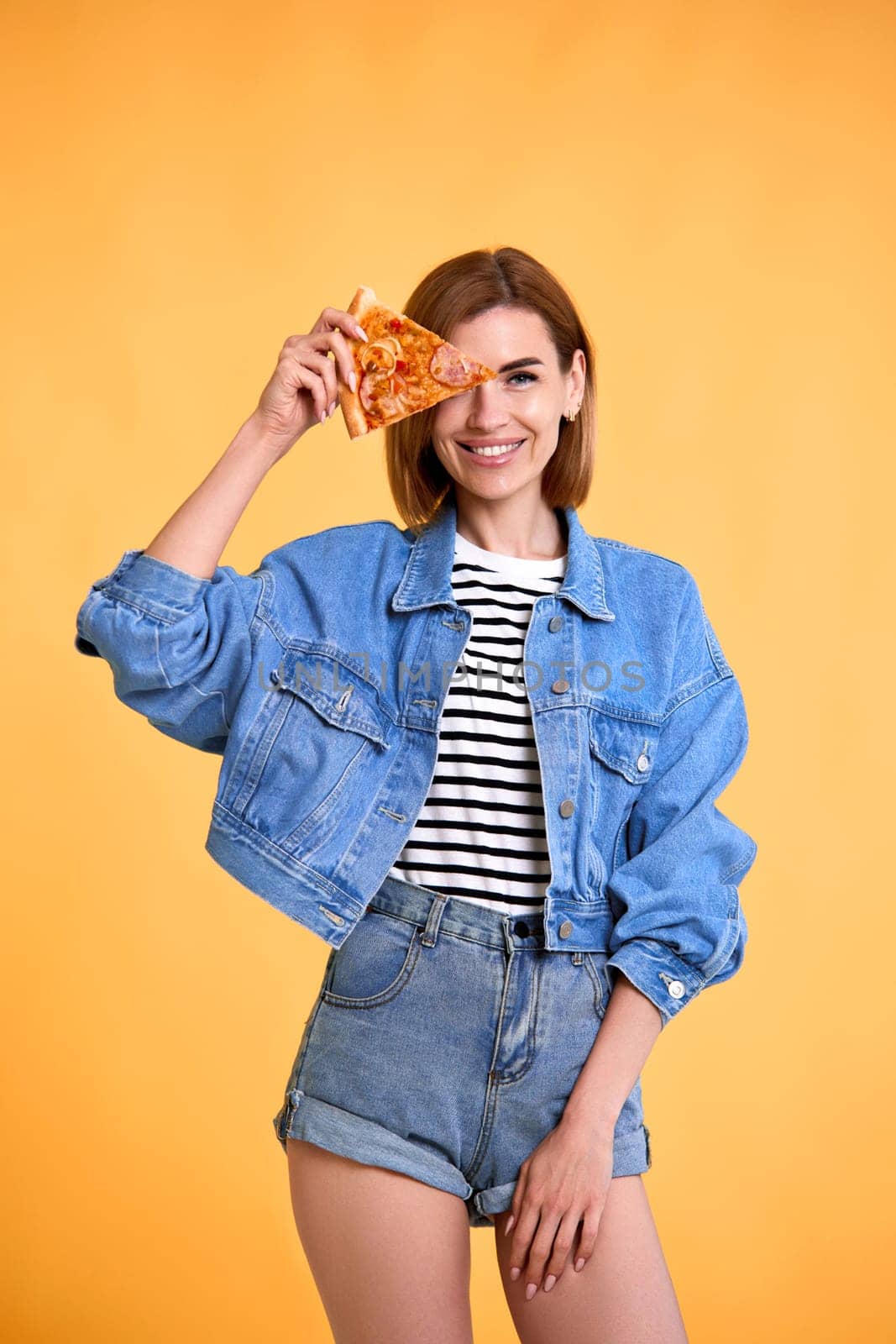 beautiful happy woman covering her eyes with a slice of fresh pizza on yellow background.