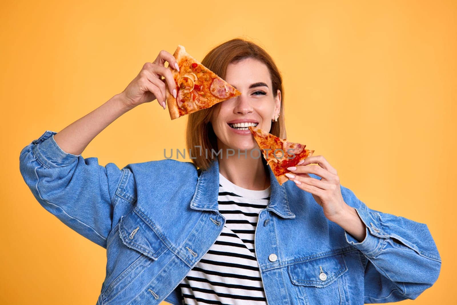 beautiful happy woman covering her eyes with slice of fresh pizza on yellow background.