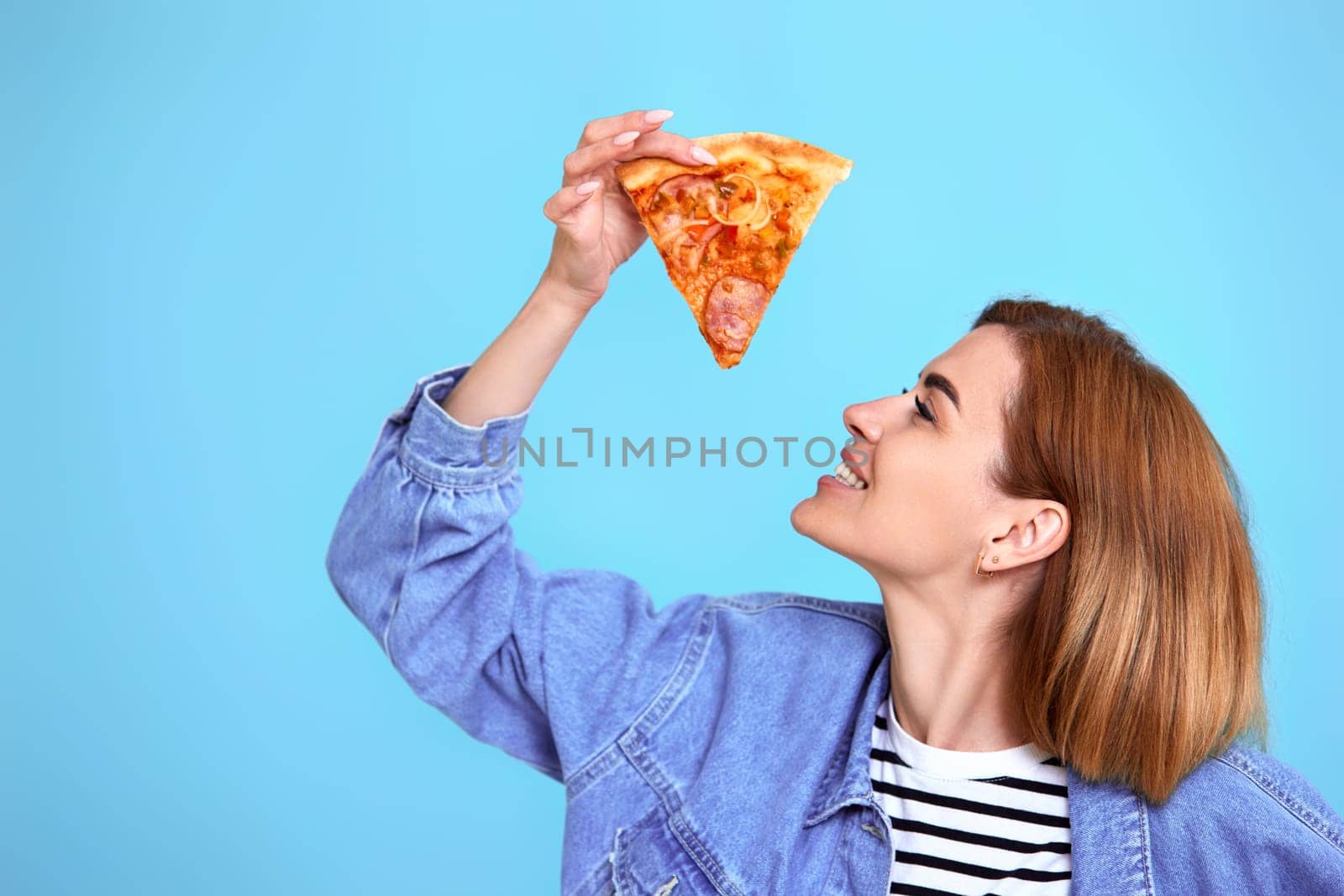 young woman eating pizza slice and looking delighted on blue background. copy space