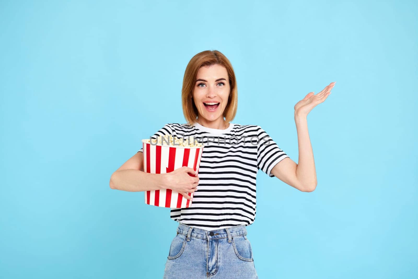smiling beautiful woman in striped t-shirt jeans denim watching film, holding bucket of popcorn isolated on blue background