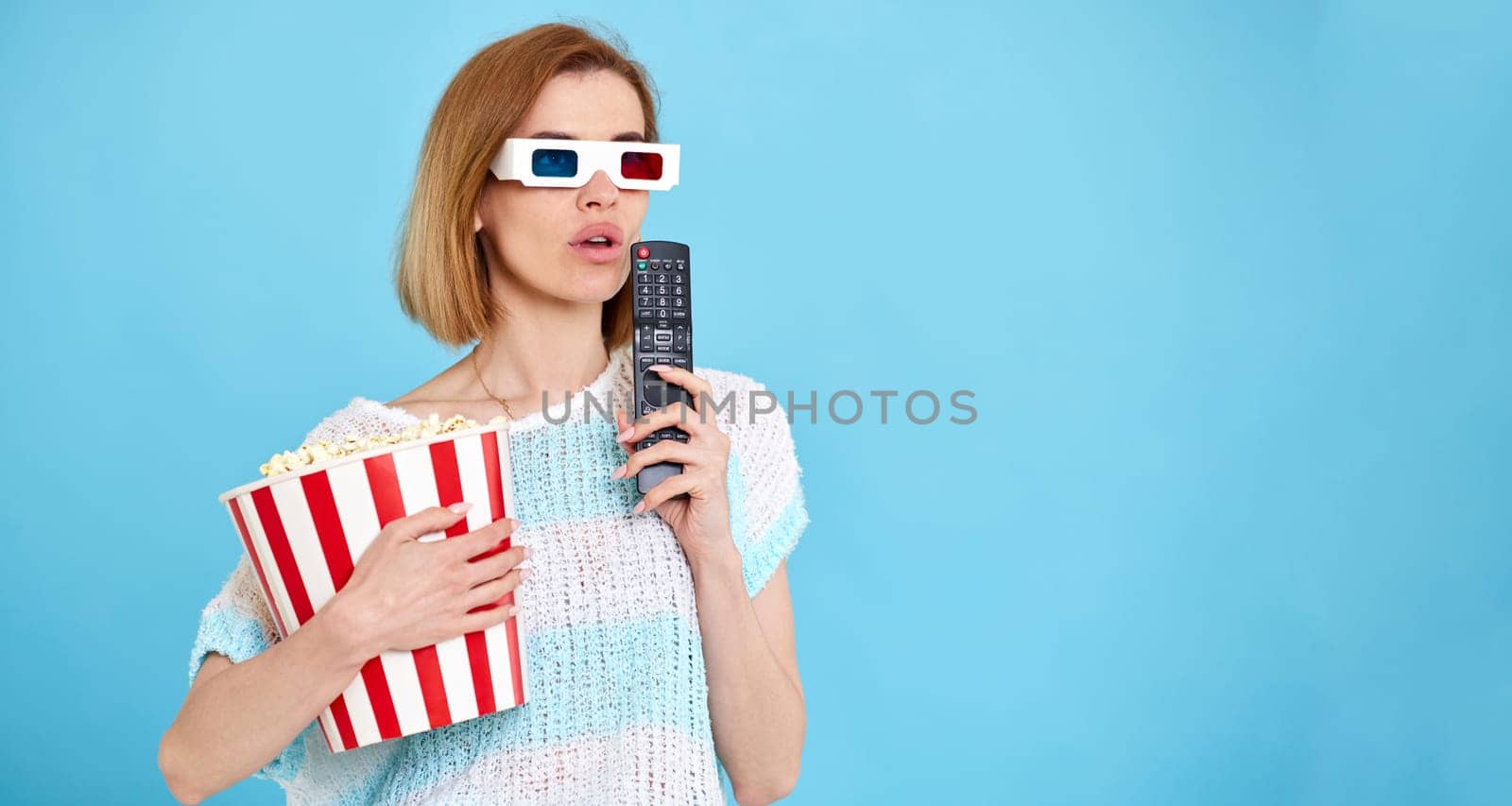 beautiful woman in 3d glasses with remote control enjoying interesting movie and holding bucket of popcorn on blue background. copy space
