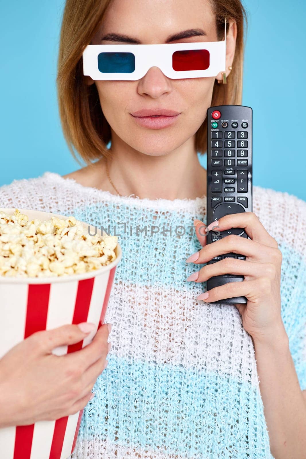 beautiful woman in 3d glasses using tv control, watching movie film and holding bucket of popcorn on blue background