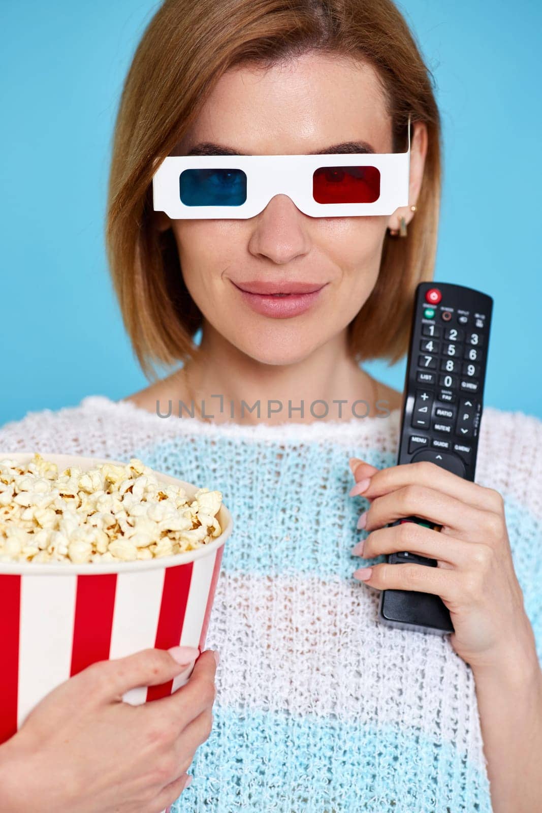 beautiful woman in 3d glasses with remote control holding bucket of popcorn on blue background