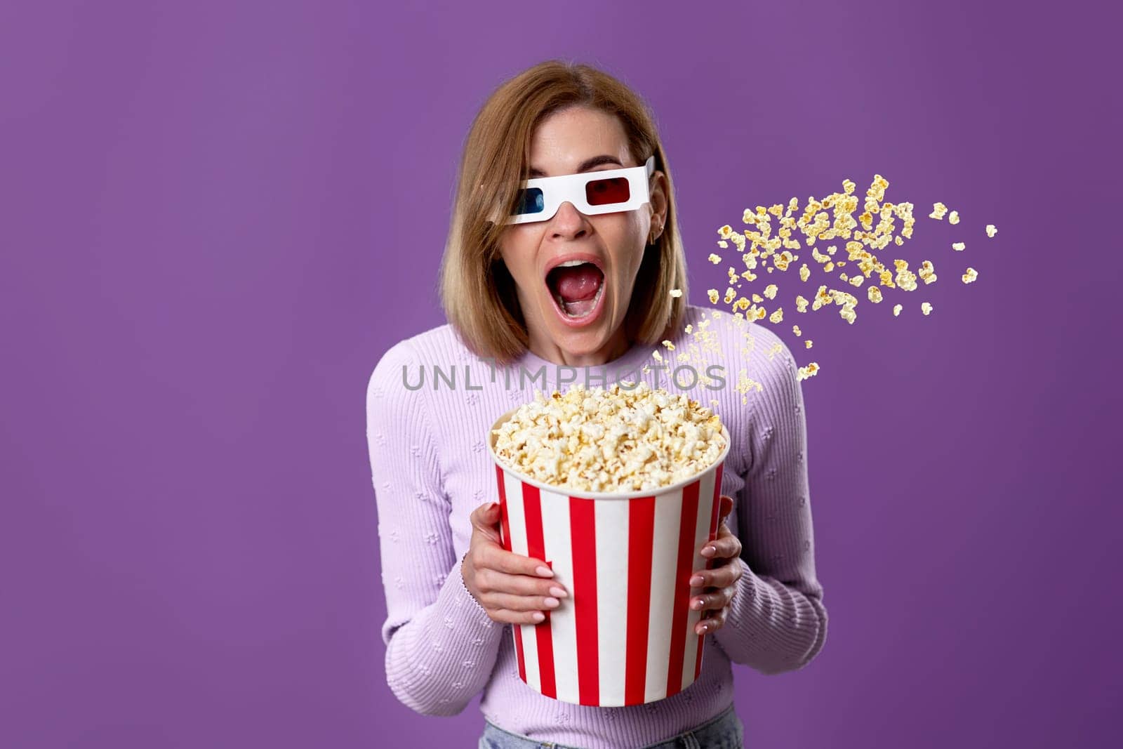 scared terrified woman in 3d glasses watching movie film, holding bucket of flying popcorn on purple background