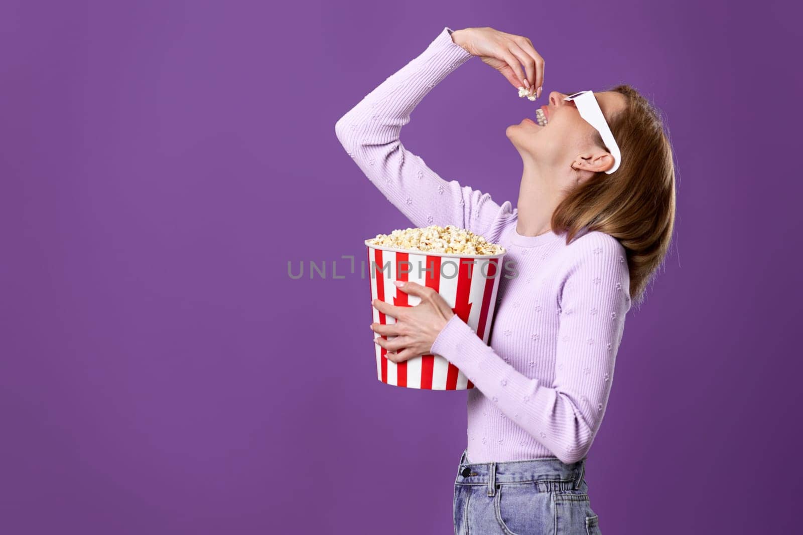 jouful caucasian woman in 3d glasses watching movie film, eating popcorn isolated on purple background