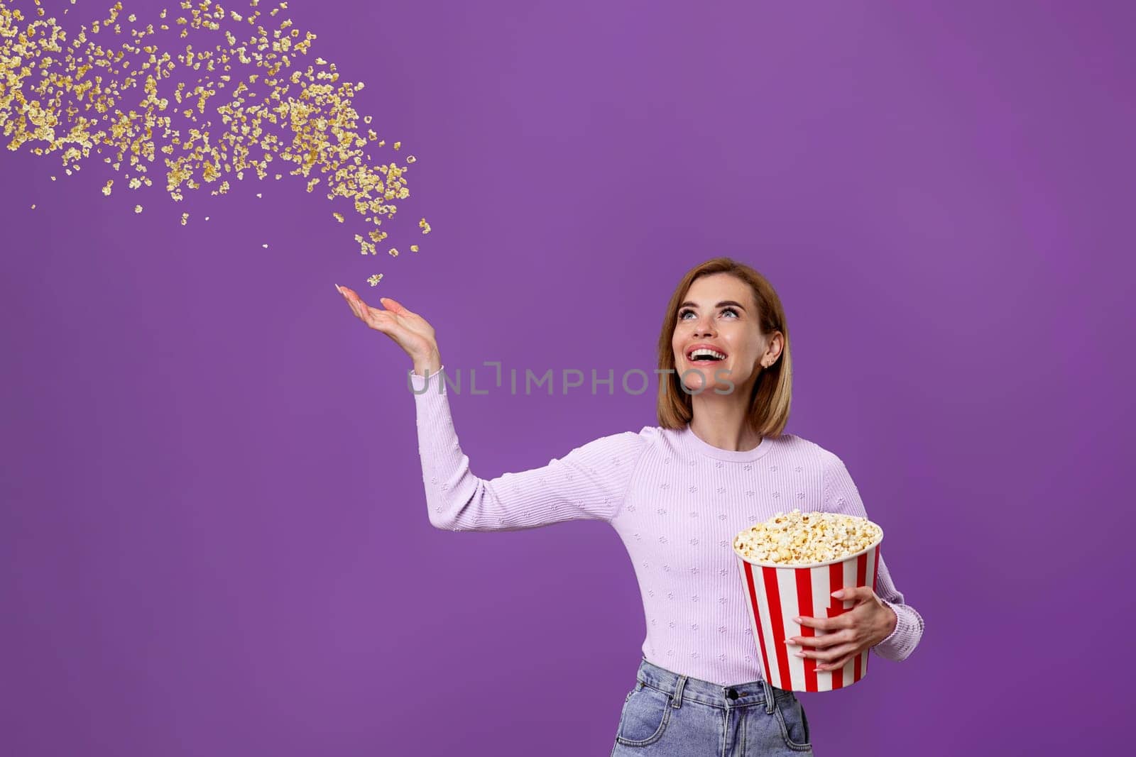 happy beautiful woman holding bucket with flying popcorn isolated on purple background