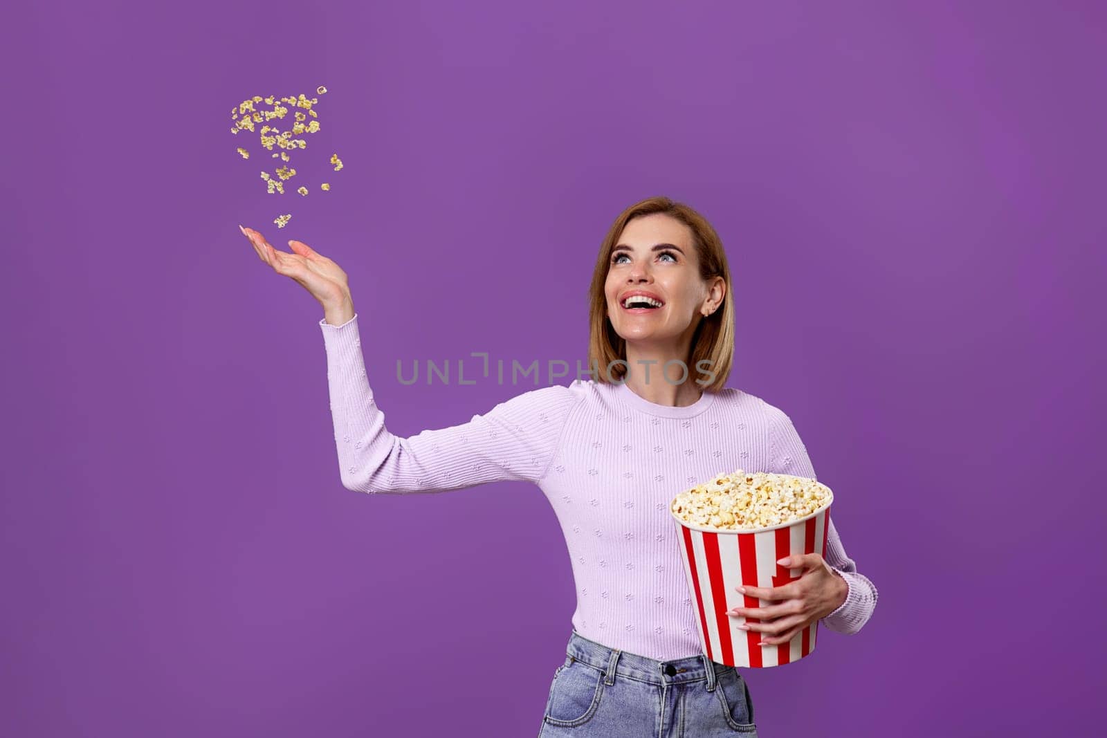 happy beautiful woman holding bucket with flying popcorn isolated on purple background