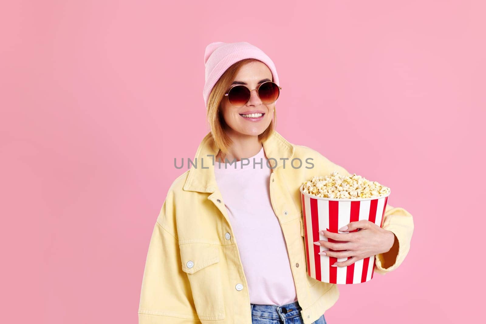 happy female hipster in sunglasses, hat watching movie film, holding bucket of popcorn isolated on pastel pink background.