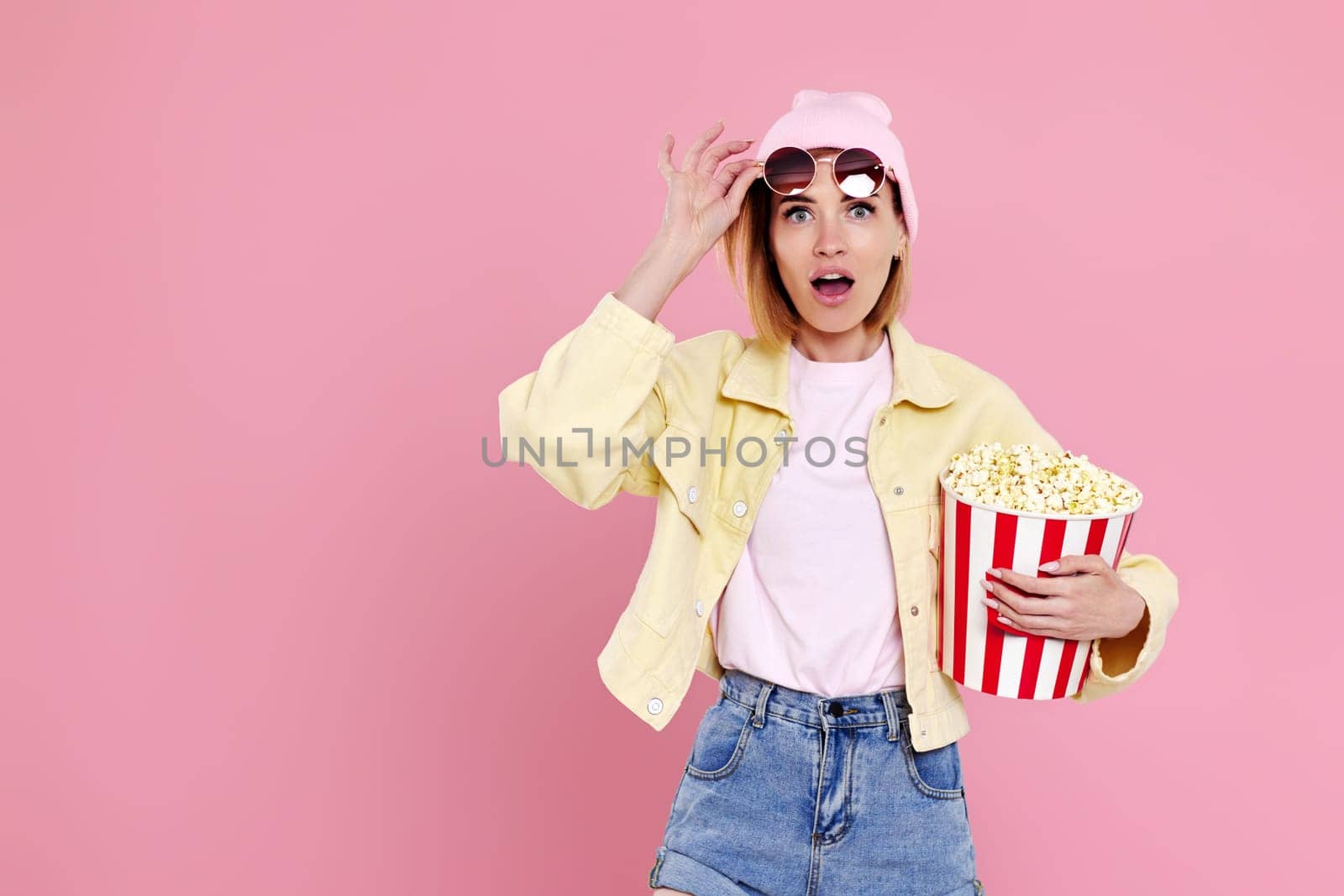surprised woman in sunglasses with bucket of popcorn watching exciting movie on pink background. copy space