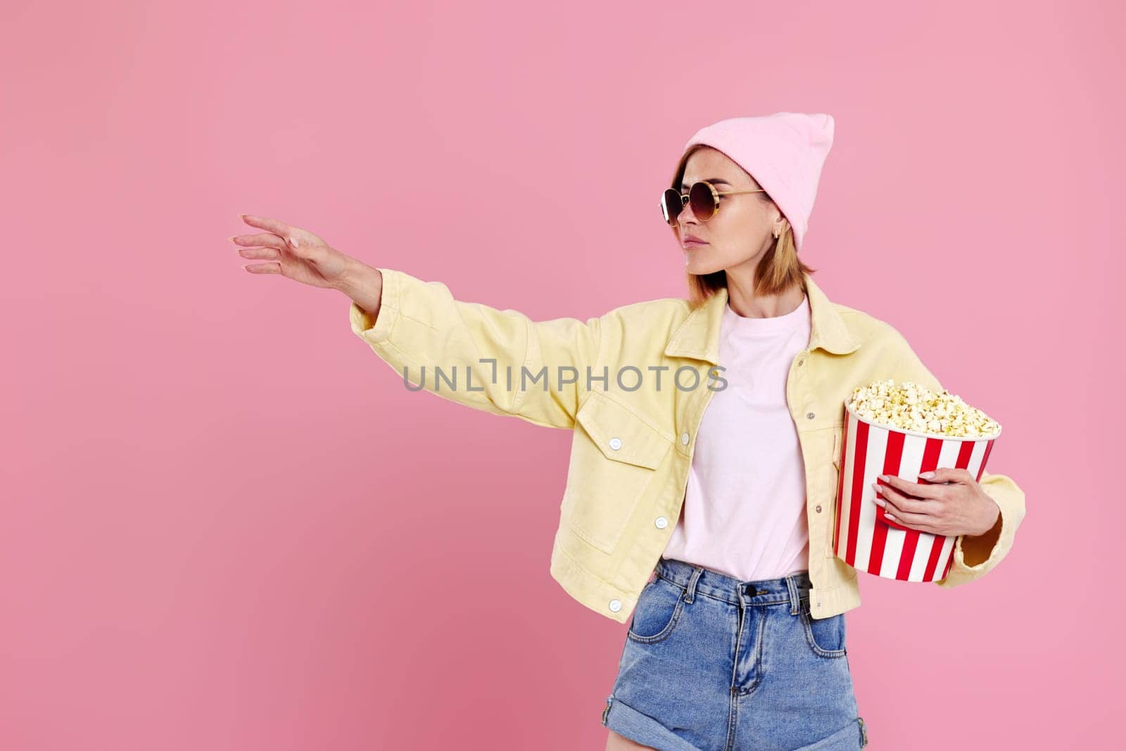 Cheerful young woman in sunglasses, hat with bucket of popcorn pointing at copy space on pastel pink background.