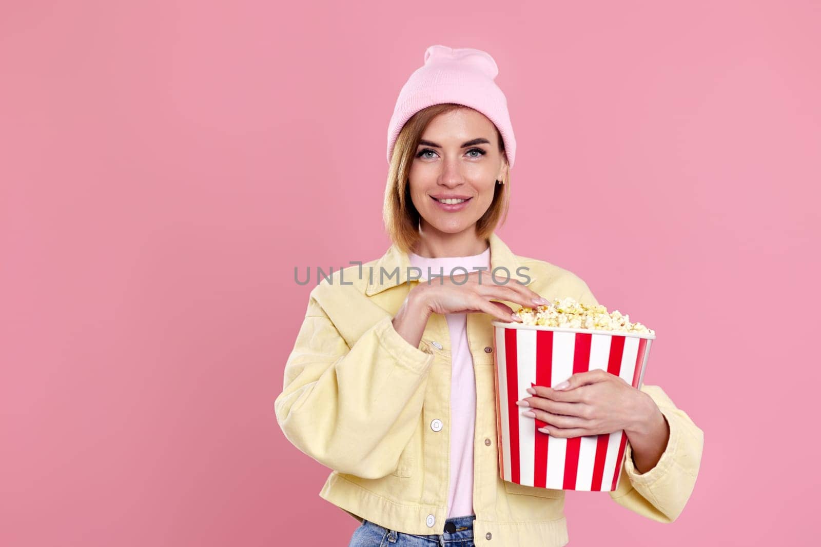 happy girl in hat watching movie film, holding bucket of popcorn isolated on pastel pink background.