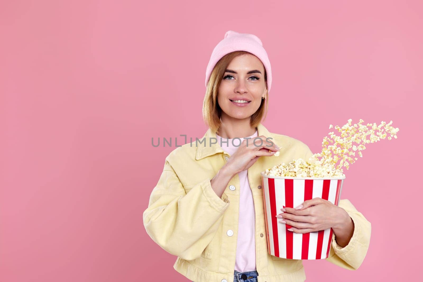 beautiful young woman in hat watching movie film, holding bucket of popcorn isolated on pastel pink background.