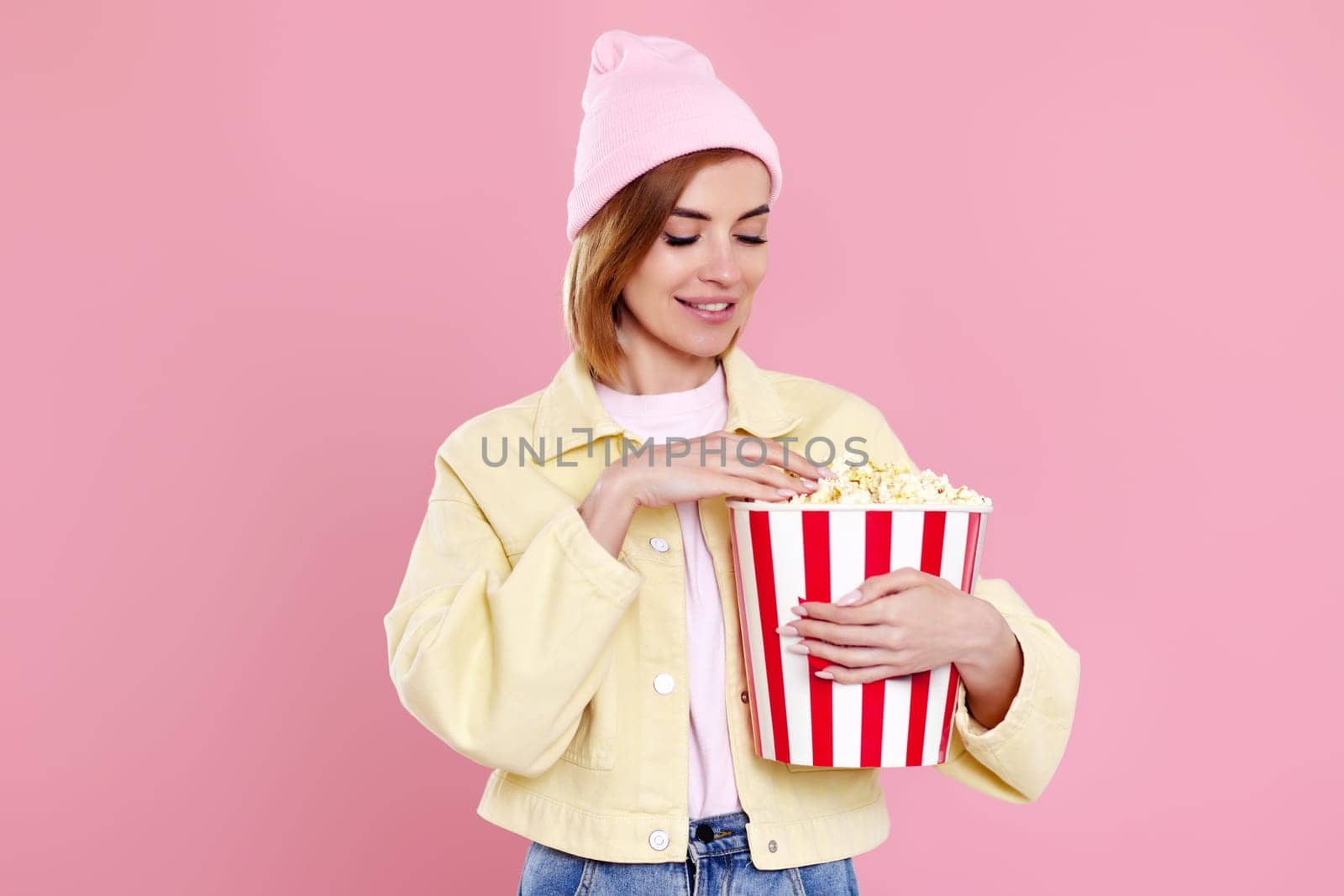 Cheerful young woman in hat watching movie film, holding bucket of popcorn on pastel pink background.