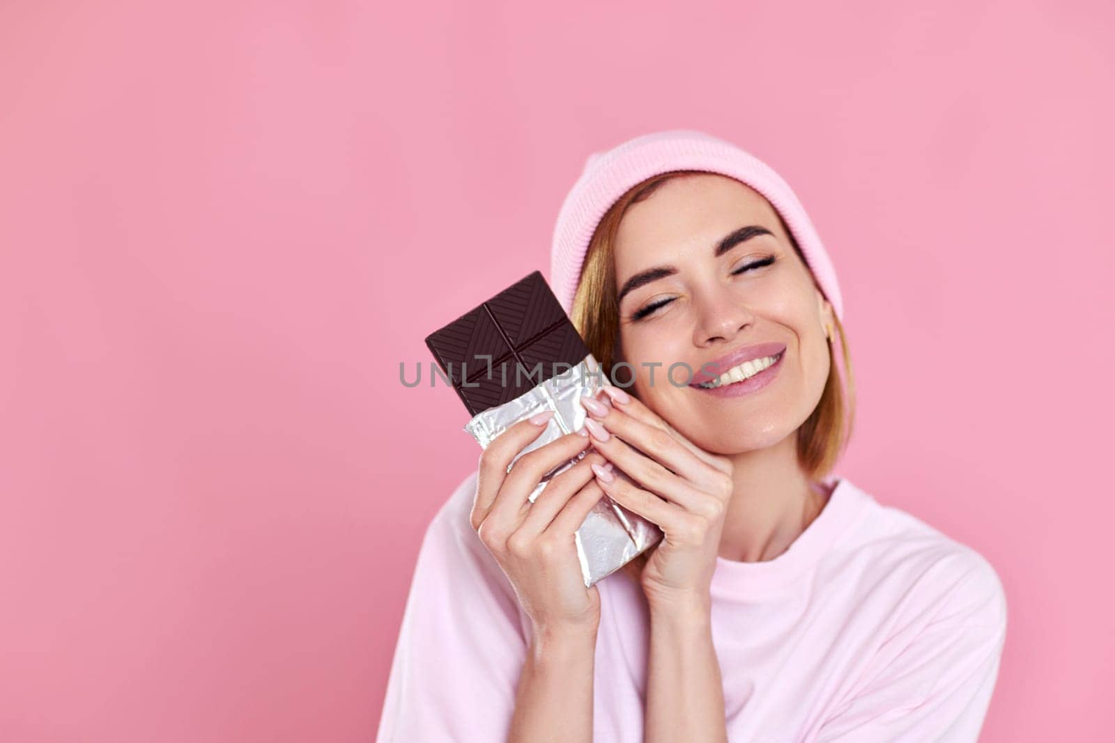 lovely smiling dreamy woman enjoying dark chocolate isolated on pastel pink background
