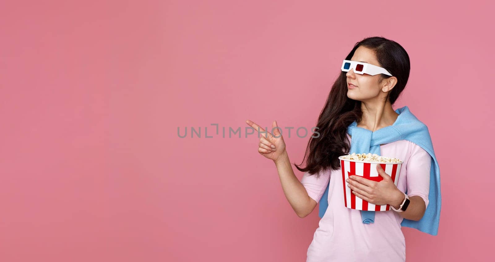 Cheerful young woman in hat with bucket of popcorn pointing at copy space on pink background.