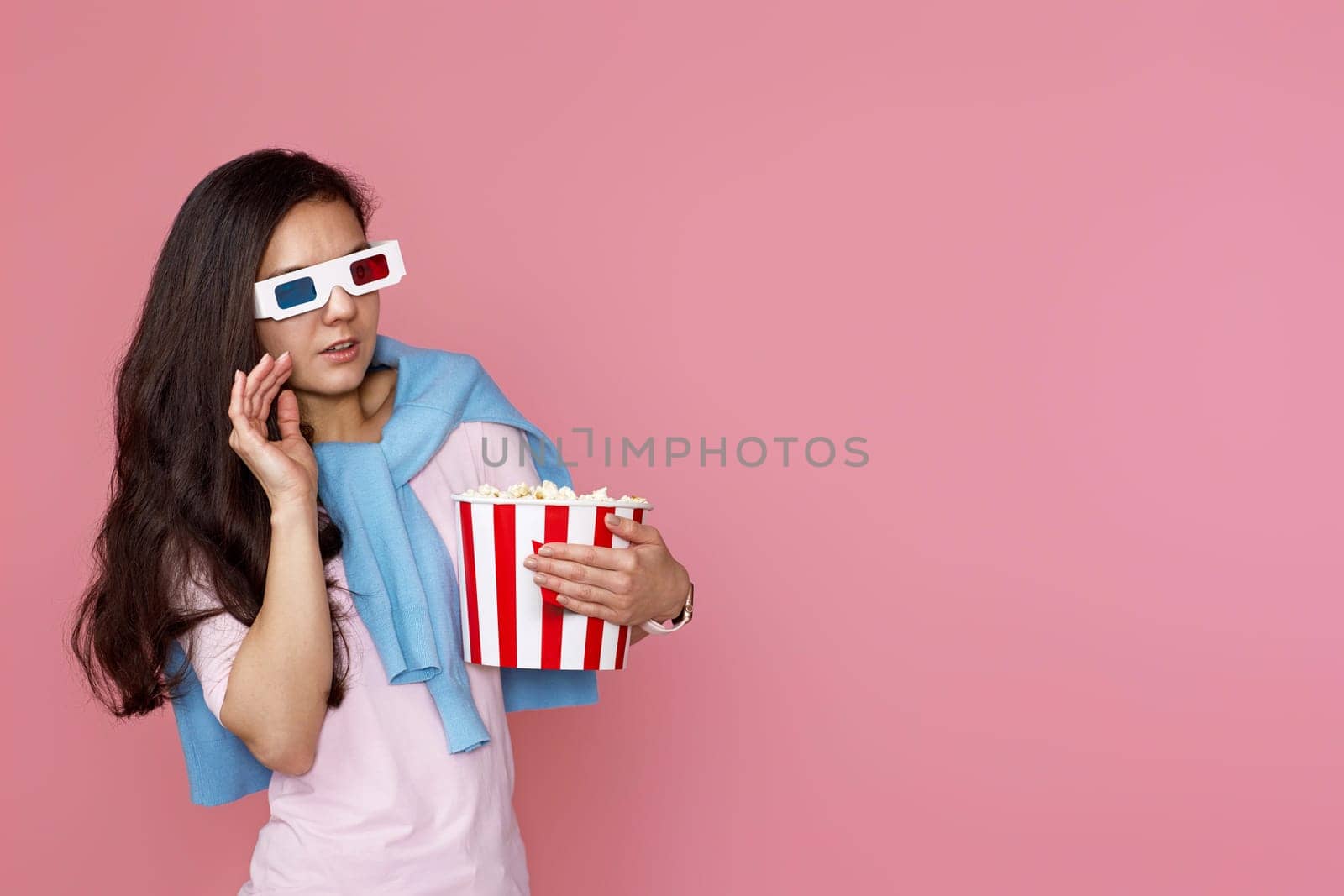 scared terrified woman watching movie film, holding bucket of popcorn on pink background. copy space