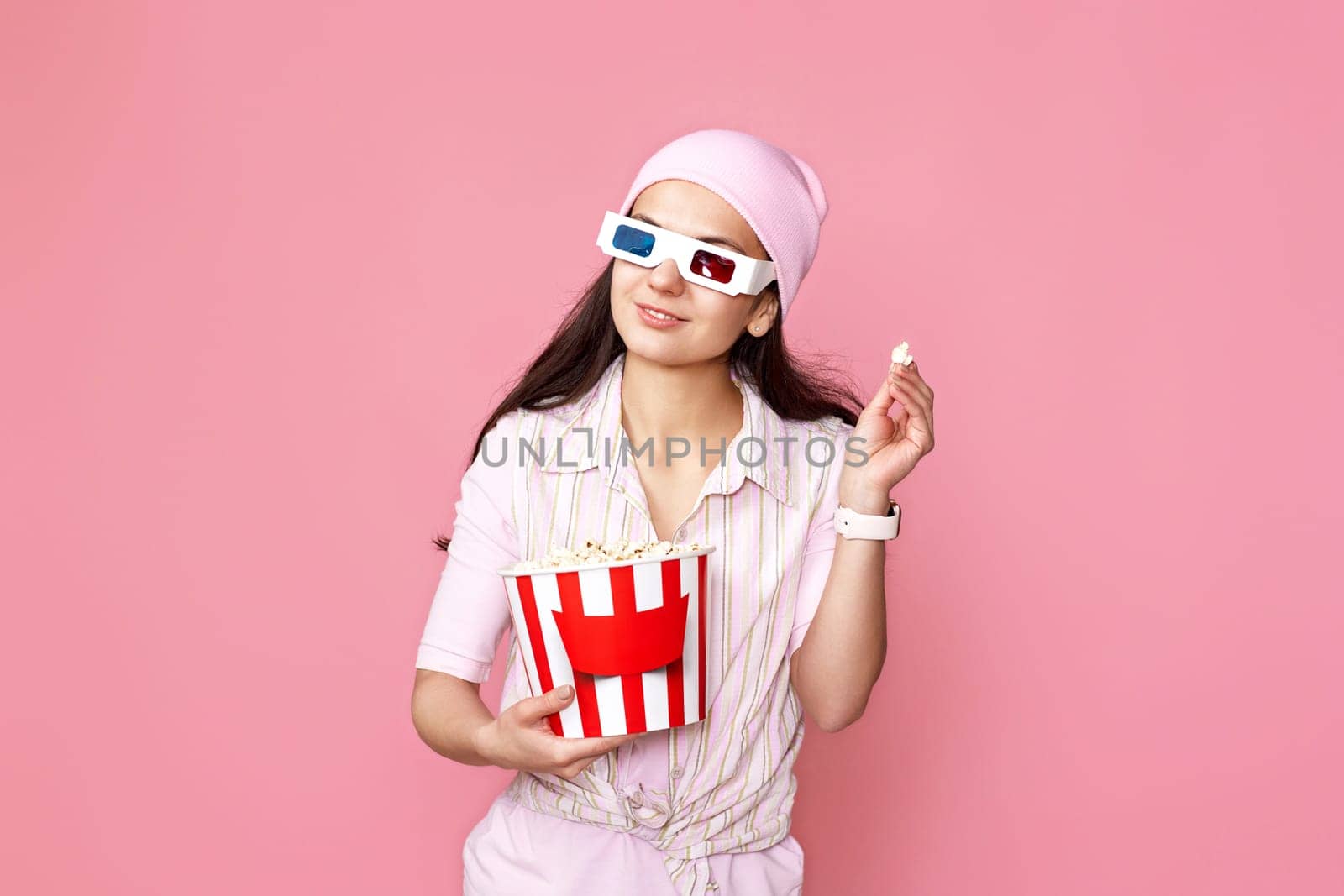 smiling caucasian girl in pink hat watching movie film, holding bucket of popcorn isolated on pastel pink background.