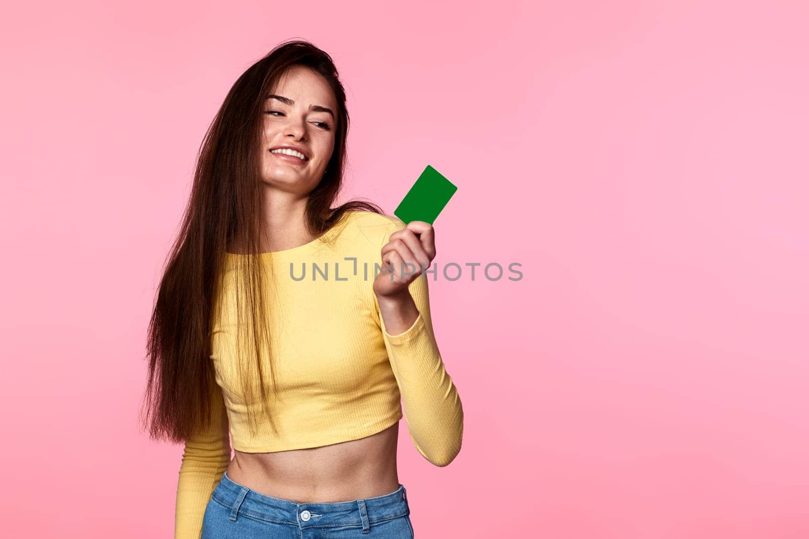 young attractive woman in yellow clothes holding plastic credit card isolated on pink background