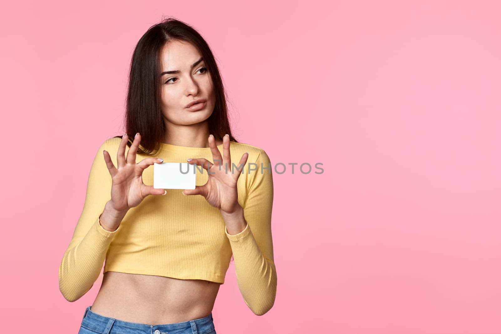 young woman in yellow clothes holding plastic credit card and looking at empty copy space isolated on pink background