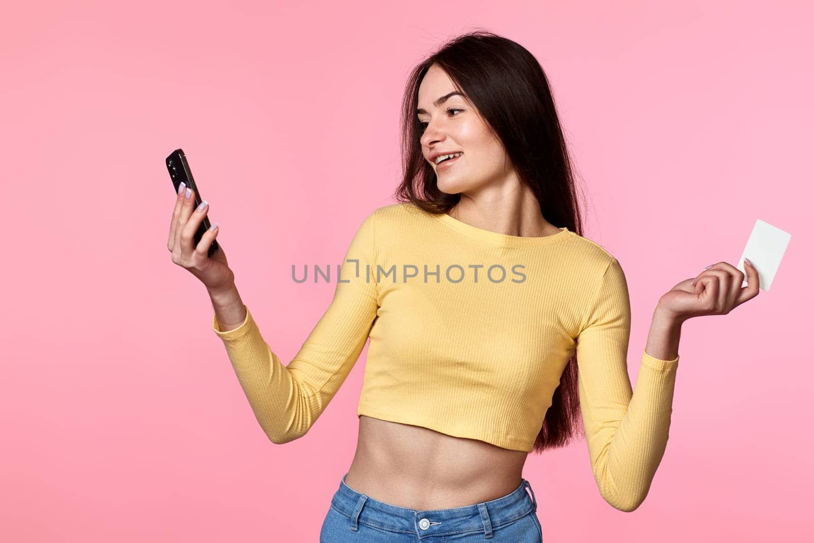 happy attractive woman in yellow clothes using mobile phone and plastic debit credit card isolated on pink background. Online payment