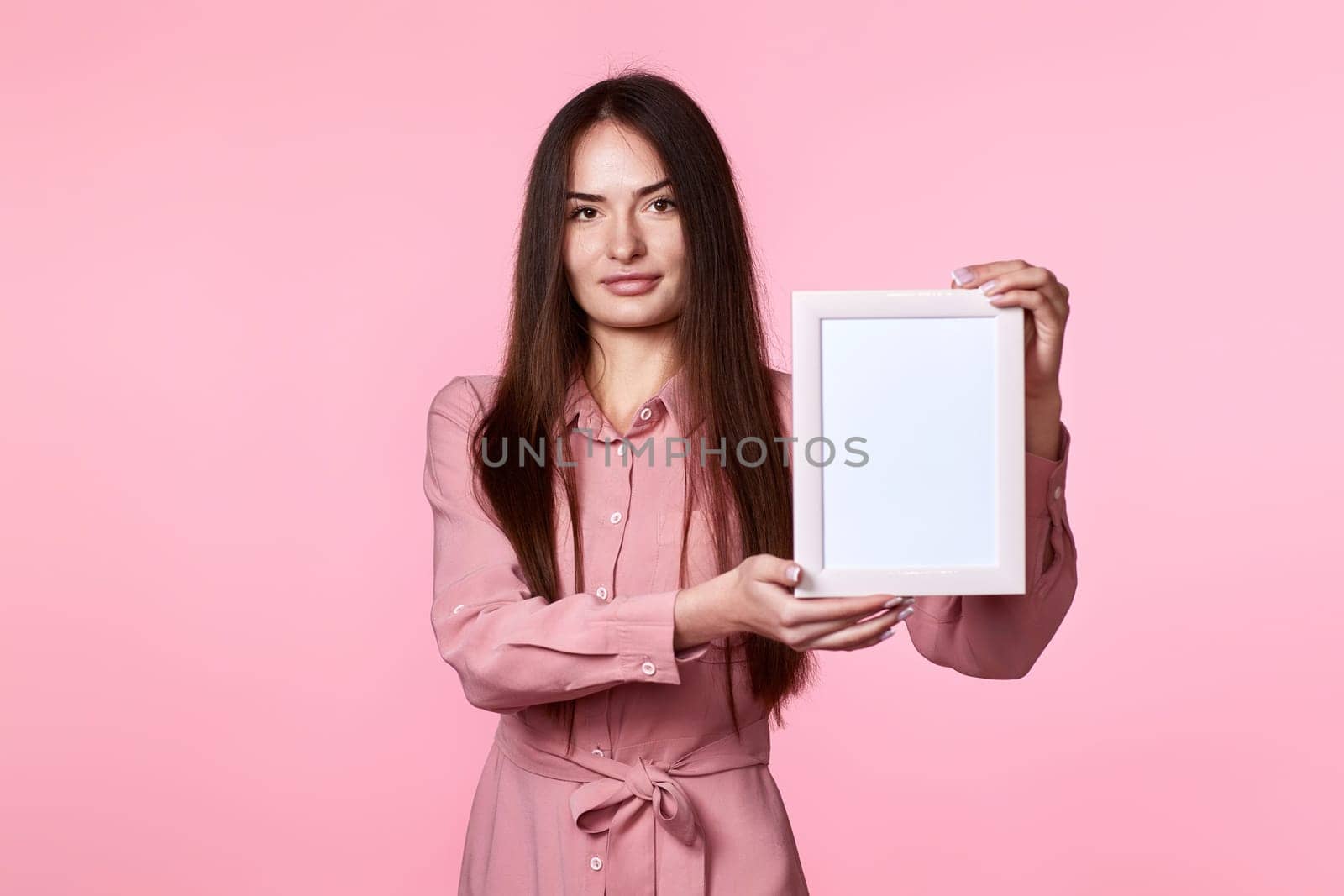 young charming woman in pink dress holding white photo frame on pink background