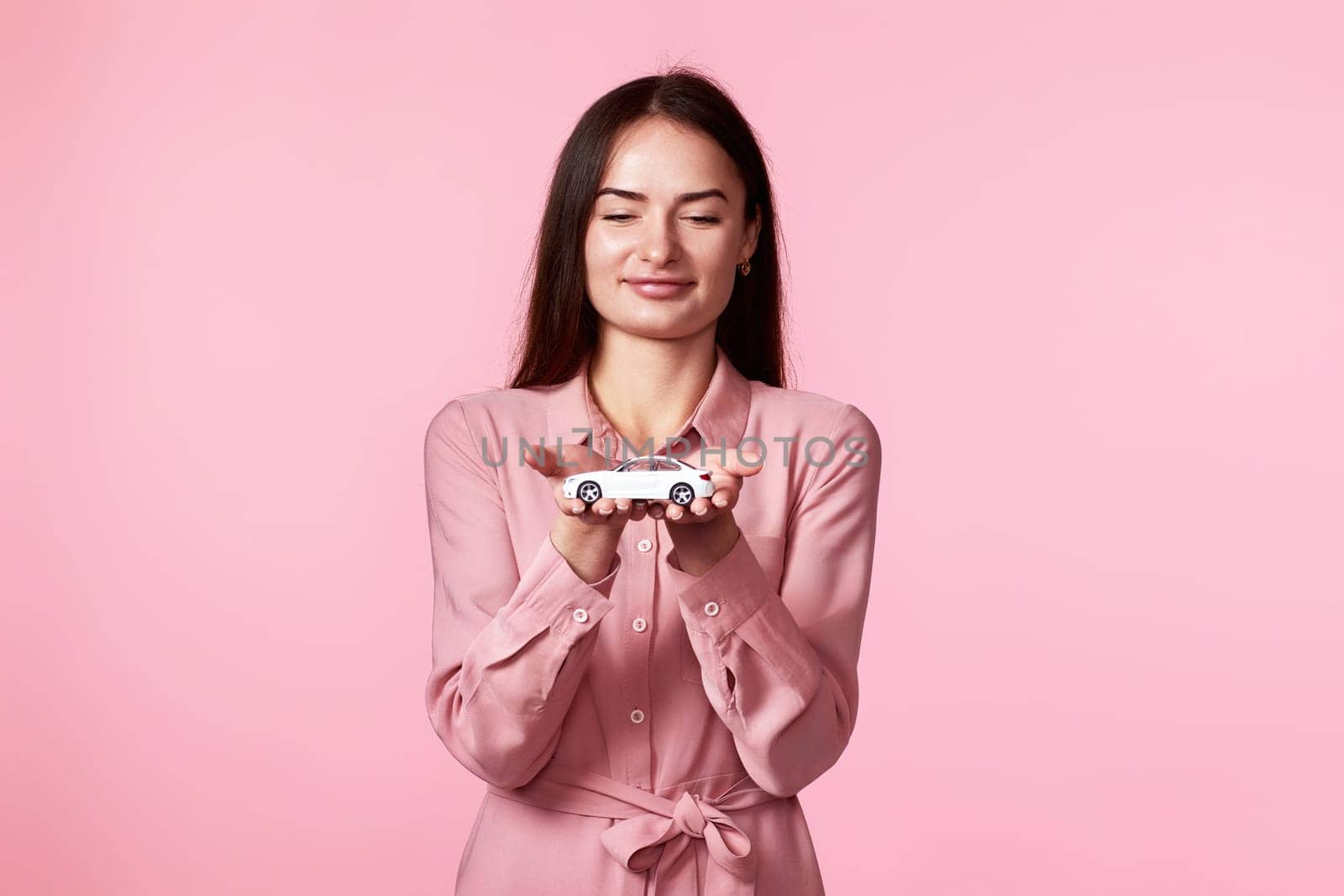 Positive woman in dress holding little white car on pink background. car protective and insurance