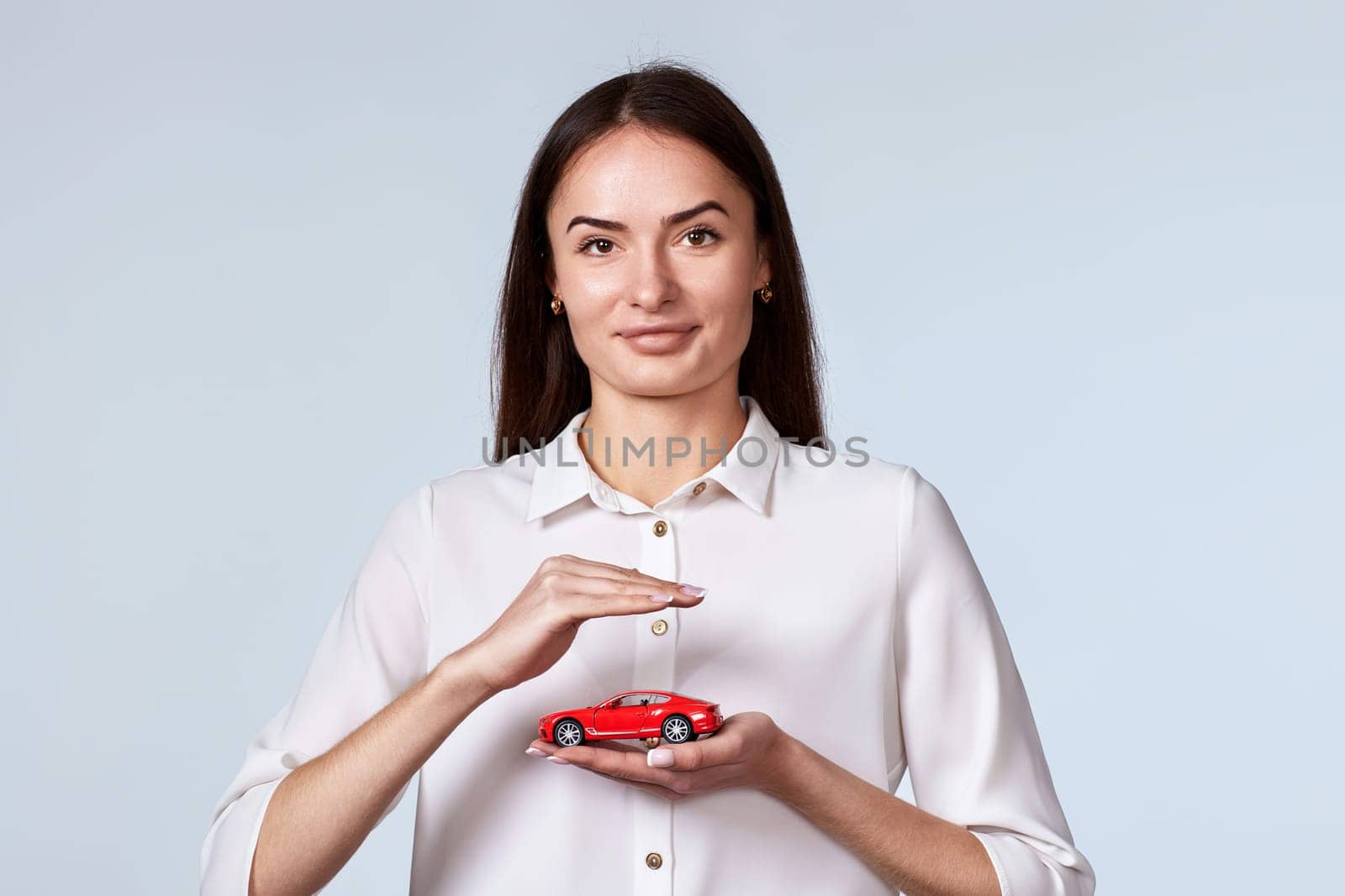 beautiful woman in white shirt holding little red car on gray background. car protective and insurance