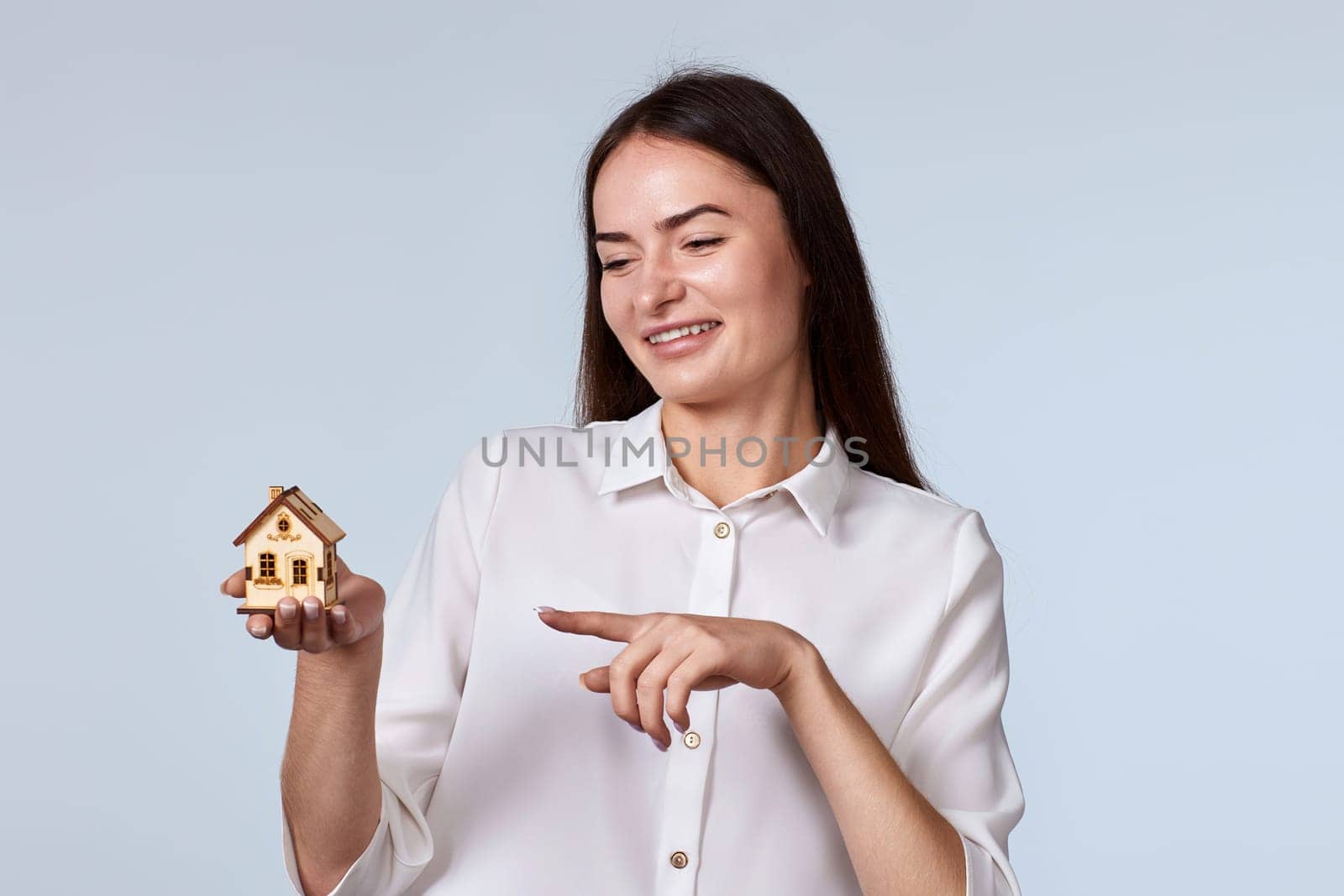 friendly smiling woman pointing finger at little house on gray background. real estate insurance concept