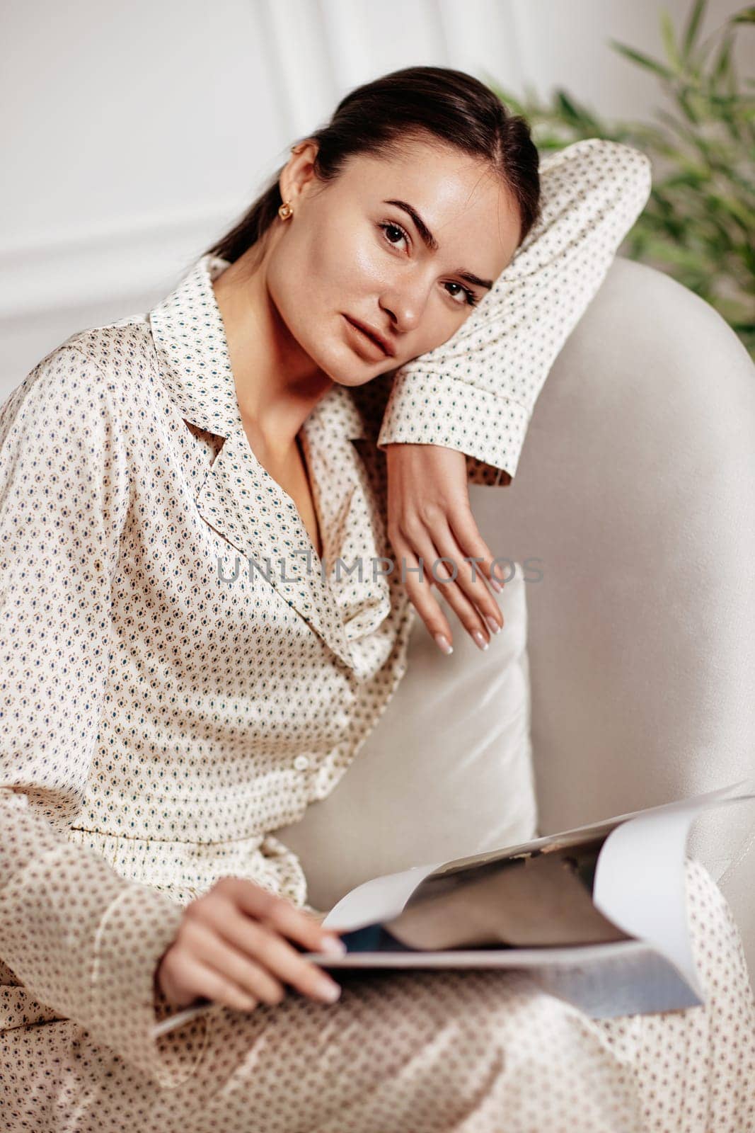 brunette woman in silk beige pajamas sits in chair and reads magazine in room