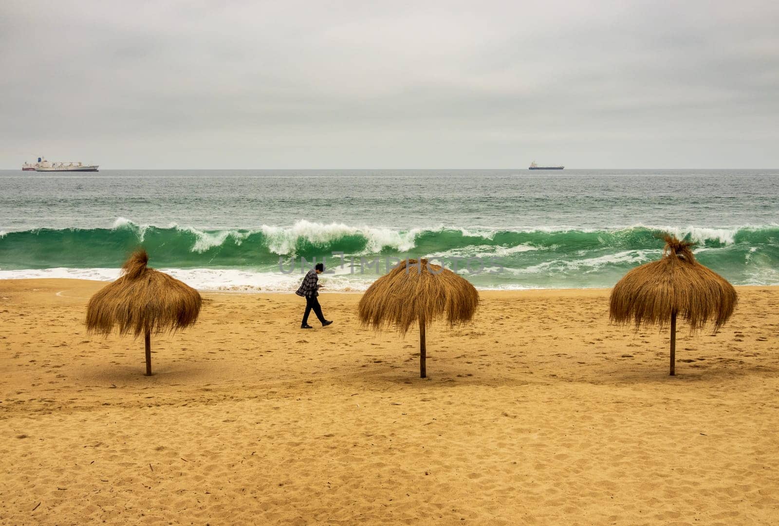 Single man walking past three palm umbrellas on an empty beach in Valparaiso Chile on a cold summer day