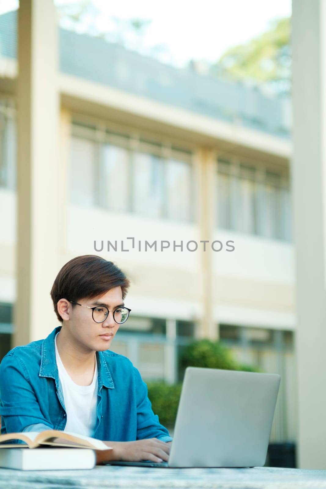 Young male college student with eyeglasses and in casual clothings sitting outdoor to study, read books, and type using laptop for school project and research. Education and E-learning concept.