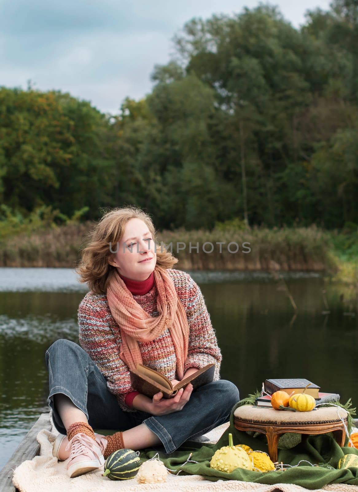 a beautiful red-haired girl reads a book on the embankment against the backdrop of autumn yellowing forests, fashion sweater. High quality photo