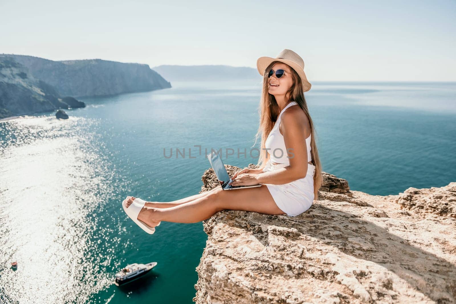 Successful business woman in yellow hat working on laptop by the sea. Pretty lady typing on computer at summer day outdoors. Freelance, travel and holidays concept.