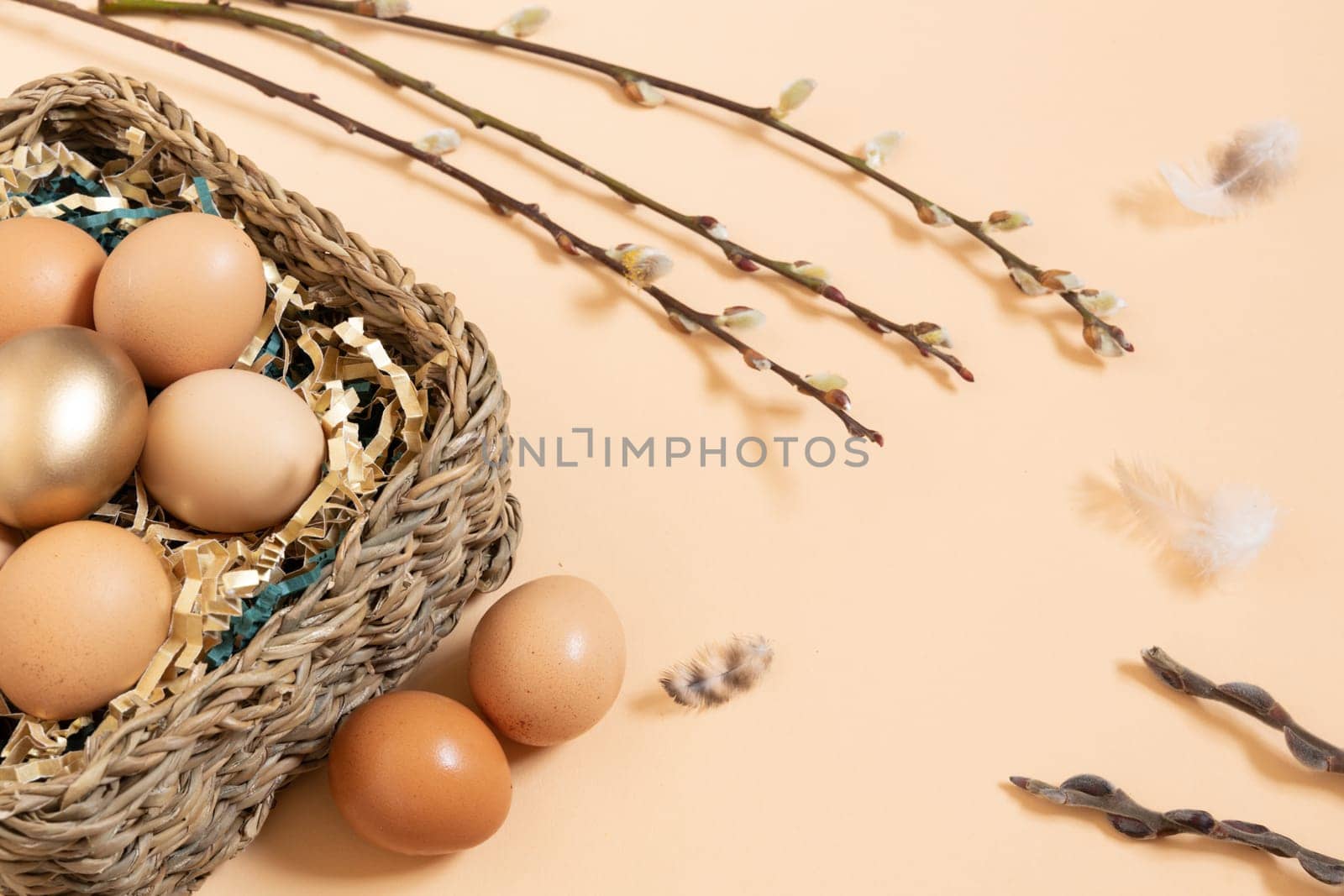 Easter eggs natural and gold in basket with paper filler . Branch of willow catkinsan feather. Light pastel orange background with copy space.