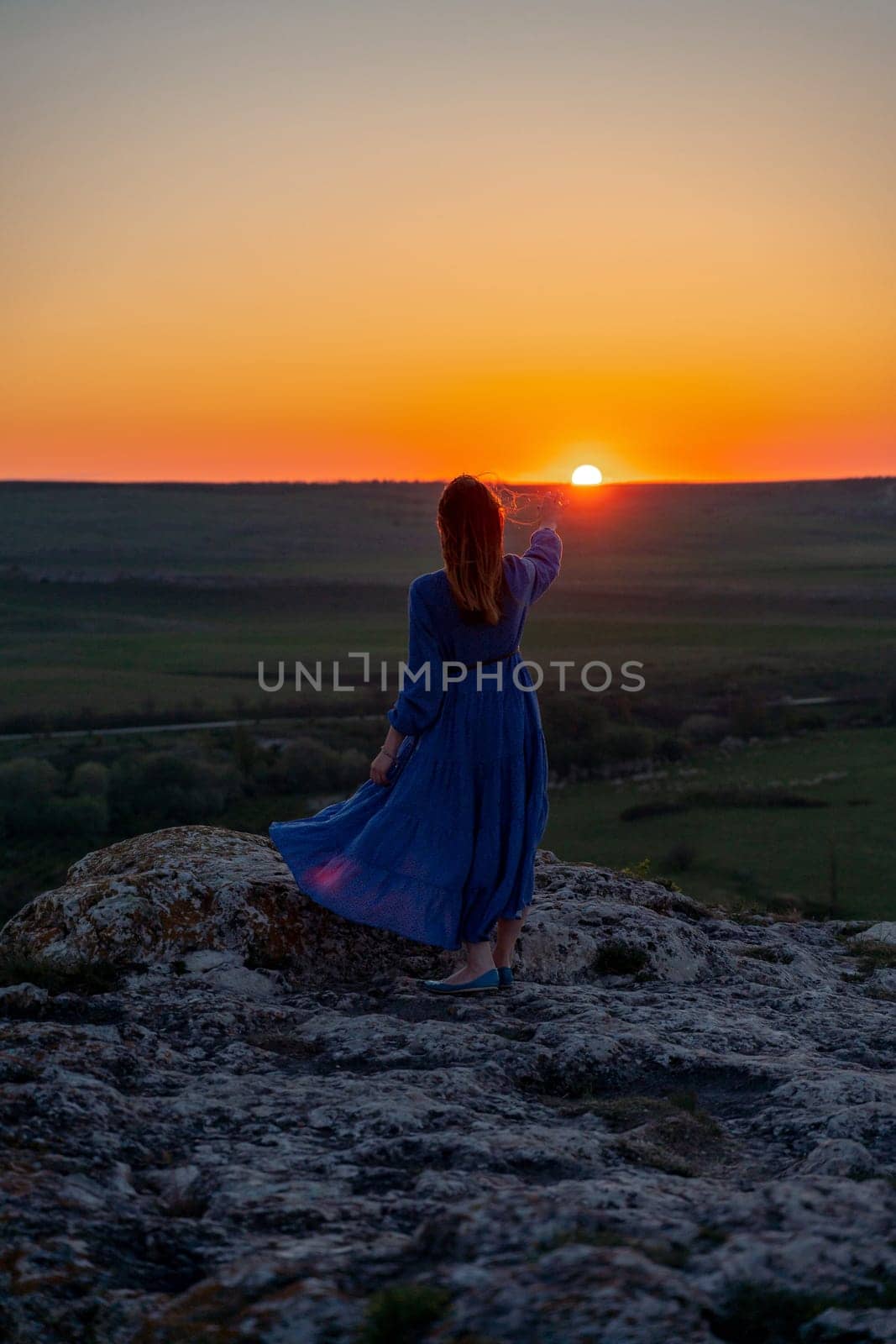 Silhouette of a girl in a blue dress standing on a rock in the mountains at sunset. She held out her hand to the sun up